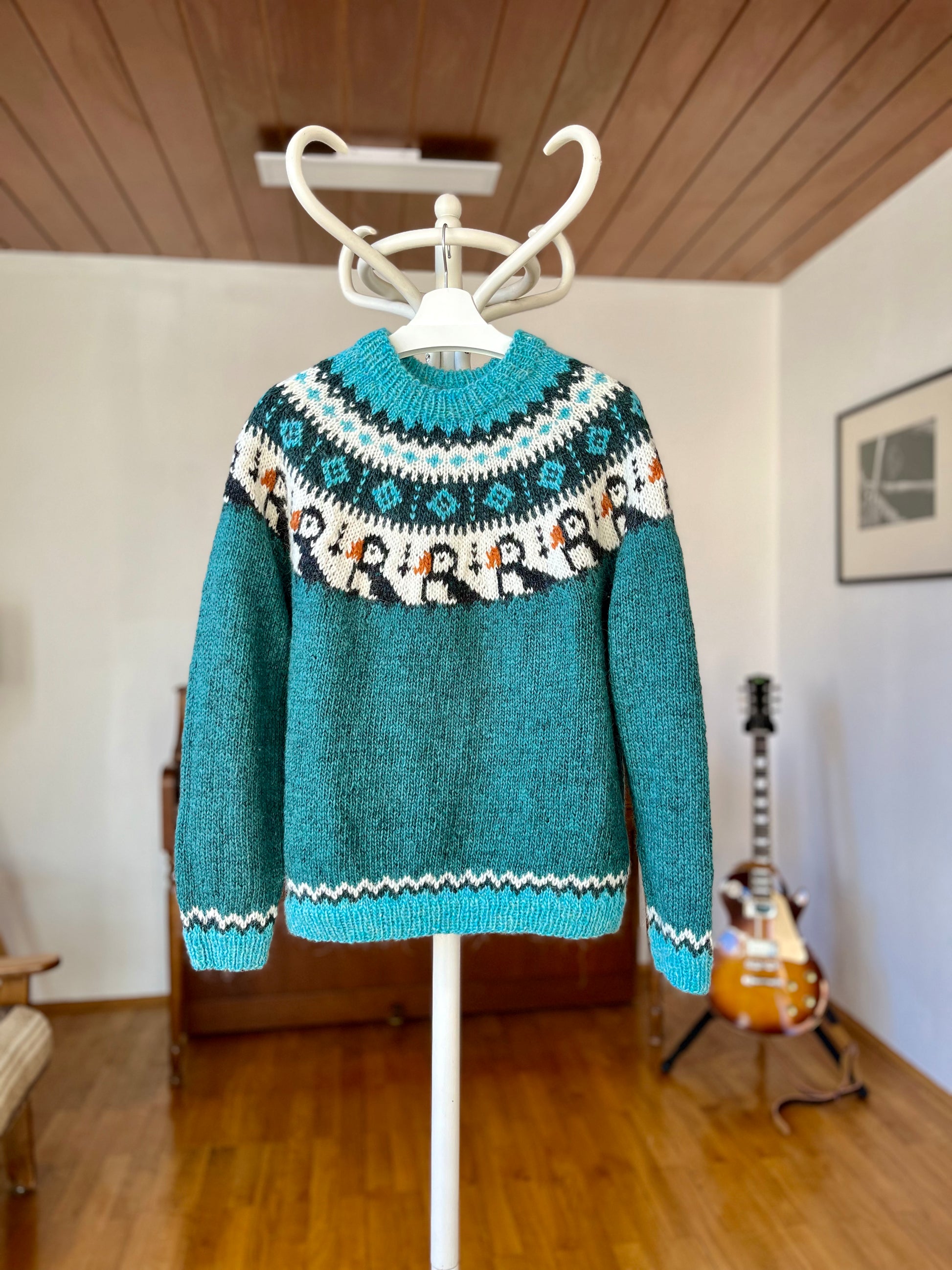 A teal knit sweater with a colorful puffin Fair Isle yoke and patterned ribbed trim, displayed on a white hanger stand in a home interior with wood floors and a guitar in the background.