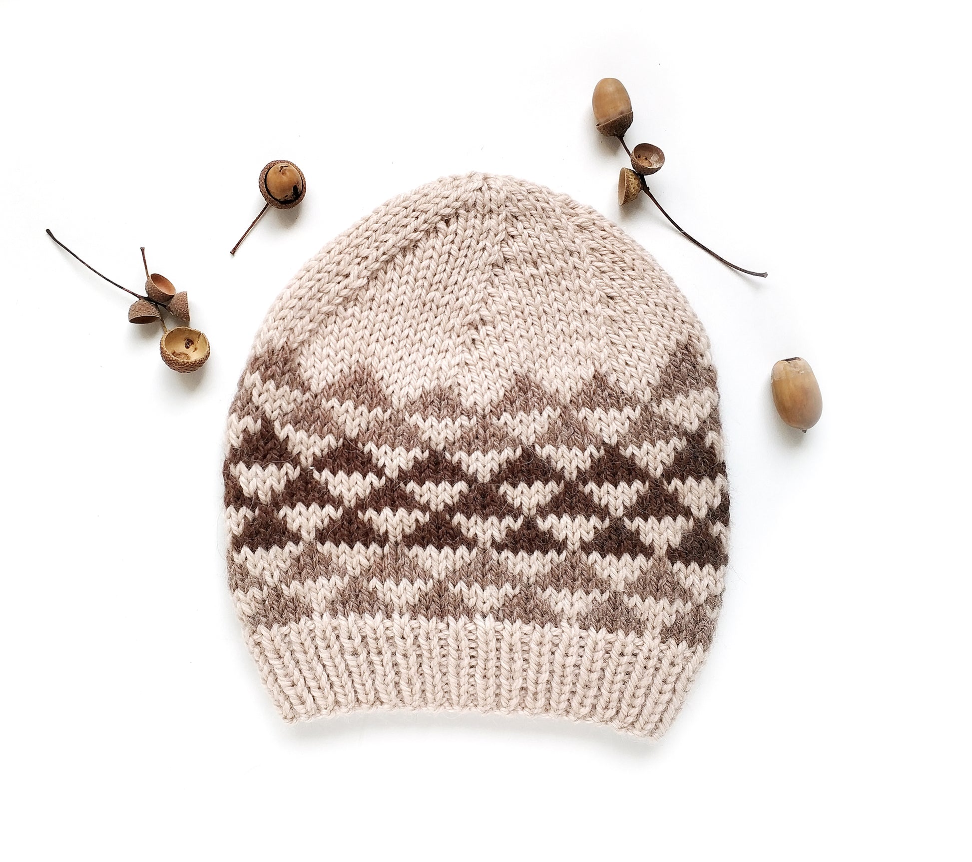 A hand-knit beanie hat for kids featuring a cream and brown Fair Isle triangles pattern, styled flat-lay with small acorns on a white background.