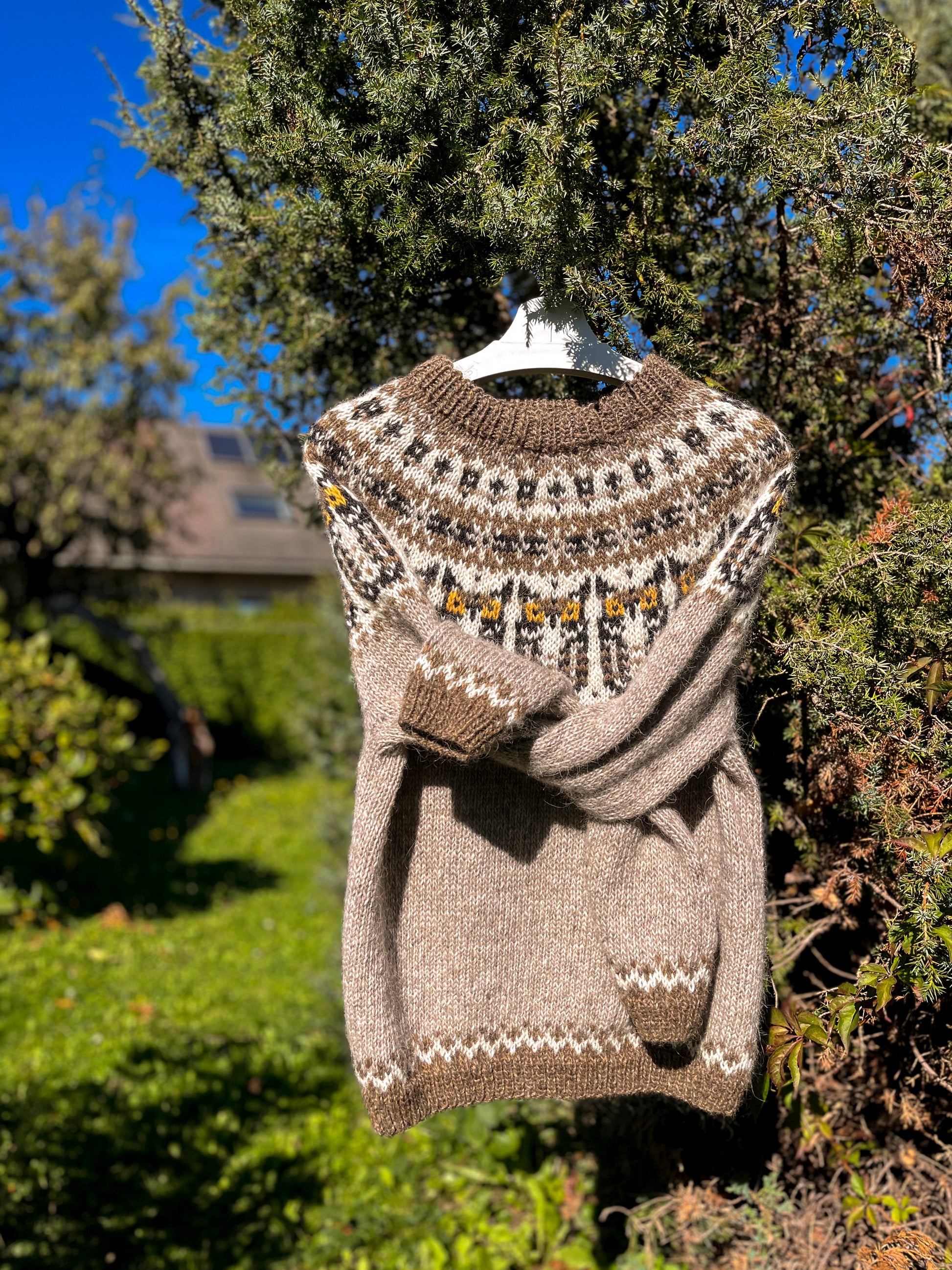 A knit pullover sweater in taupe and brown with a Fair Isle yoke pattern, hanging on a white hanger outdoors against green bushes and trees.