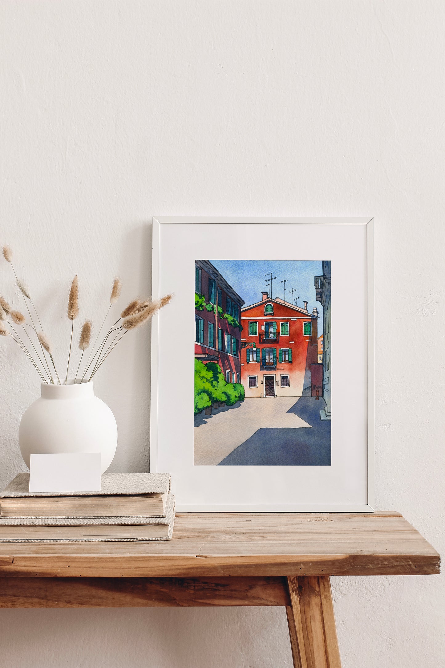 Framed watercolor artwork of a Venetian street scene on a wooden table with a vase and books.