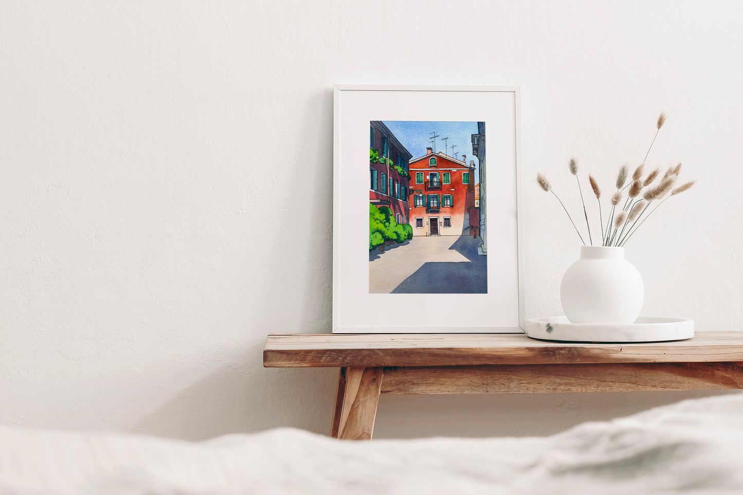 Framed watercolor and ink artwork of a Venetian street scene on a wooden shelf with a vase of dried plants.