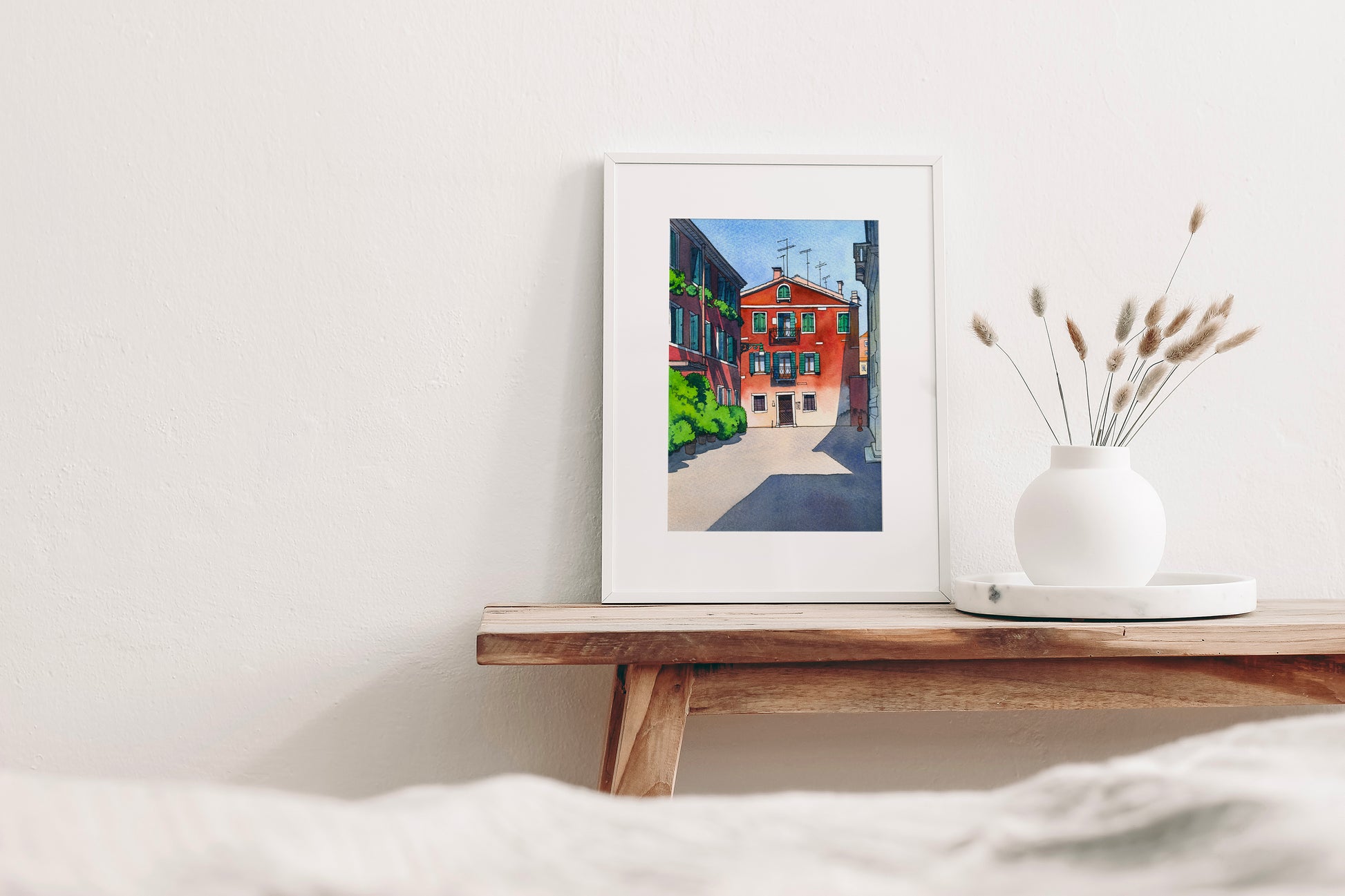 Framed watercolor and ink artwork of a Venetian street scene on a wooden shelf with a vase of dried plants.