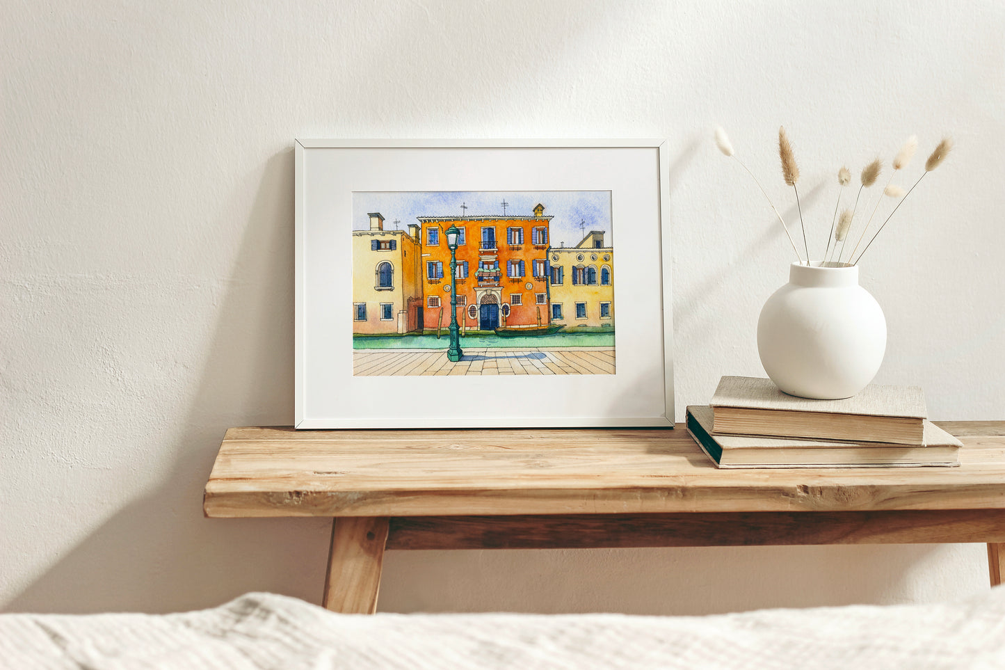 Framed watercolor artwork of a colorful building in Venice, Italy on a wooden shelf with a vase and books.