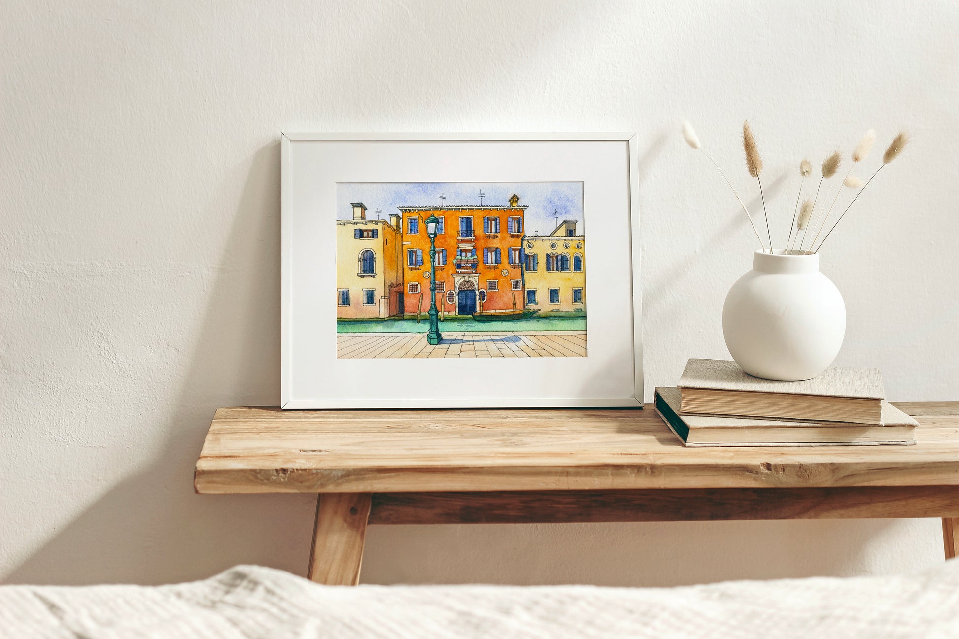 Framed watercolor artwork of a colorful building in Venice, Italy on a wooden shelf with a vase and books.