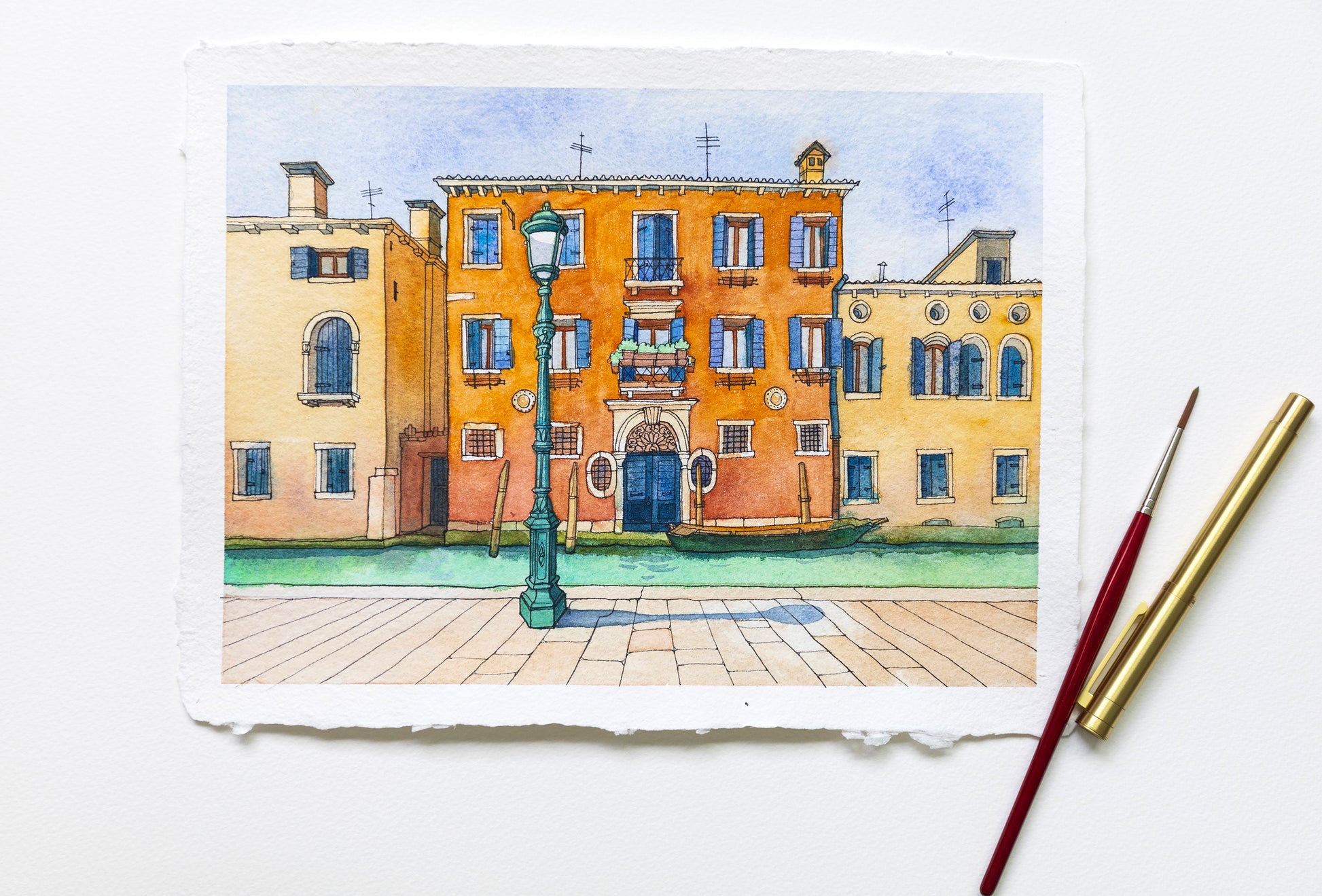 Watercolor painting of a street scene with buildings in Venice and a lamp post, accompanied by two paintbrushes.