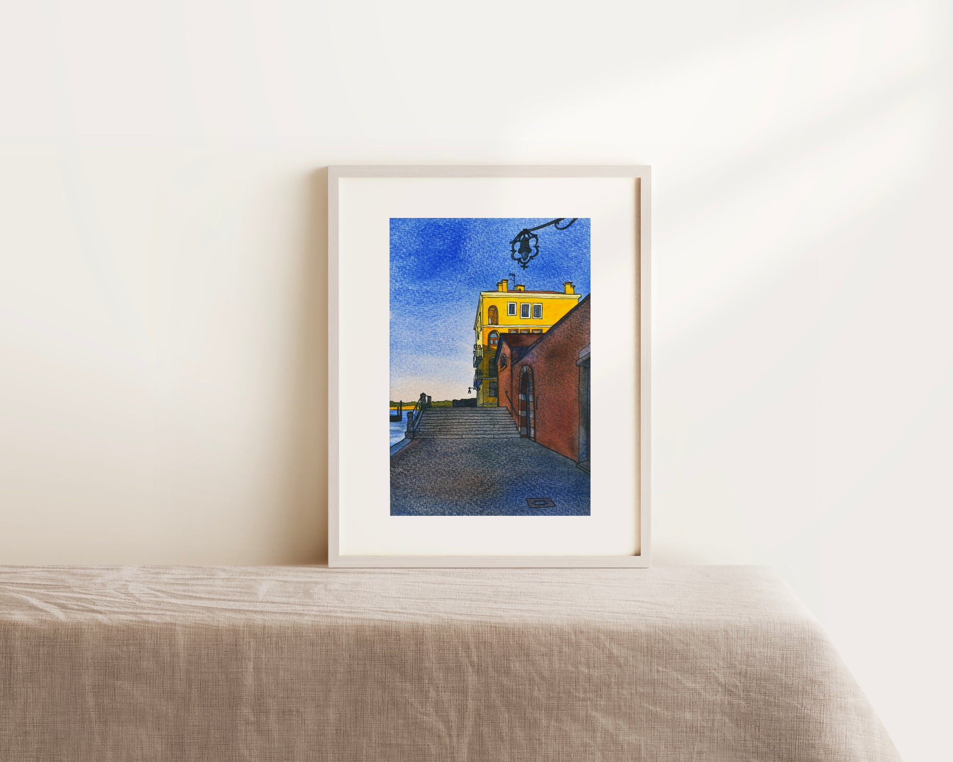 Framed watercolor and ink artwork of a yellow building on a white wall above a table.