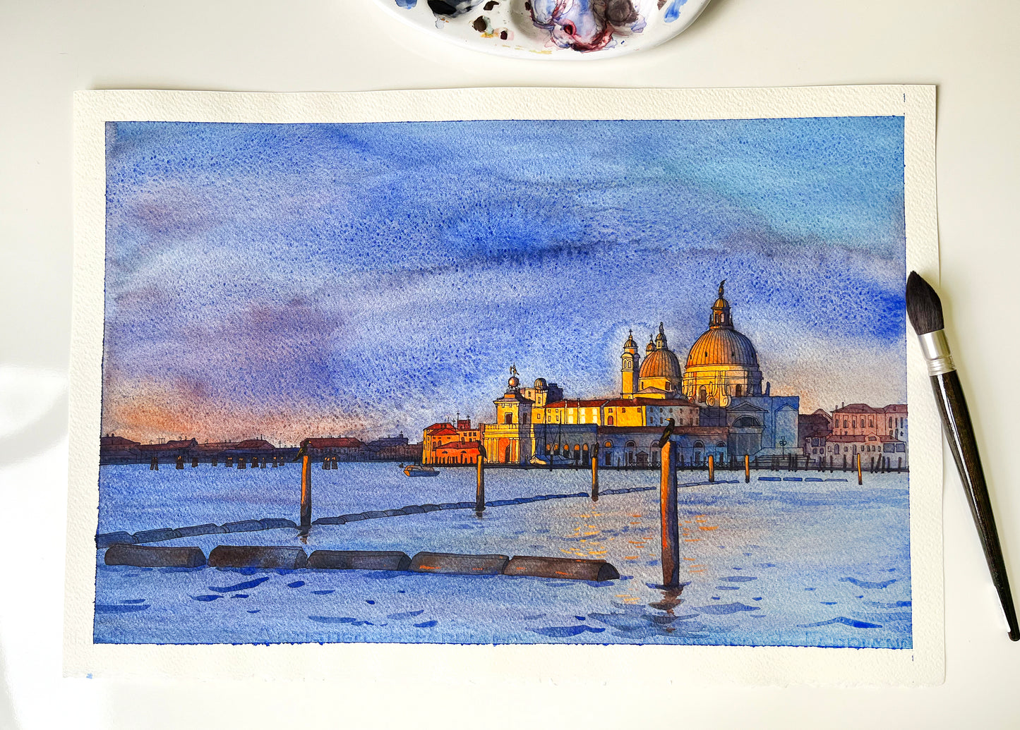 Watercolor painting of Venice San Marco basin at sunrise with illuminated buildings on a white surface.