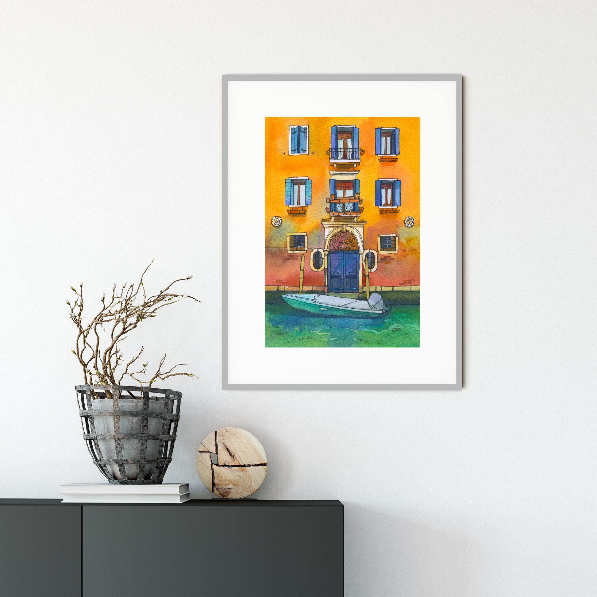 Framed watercolor artwork of a Venetian canal scene with a yellow building and blue door, placed on a white wall above a dark surface.