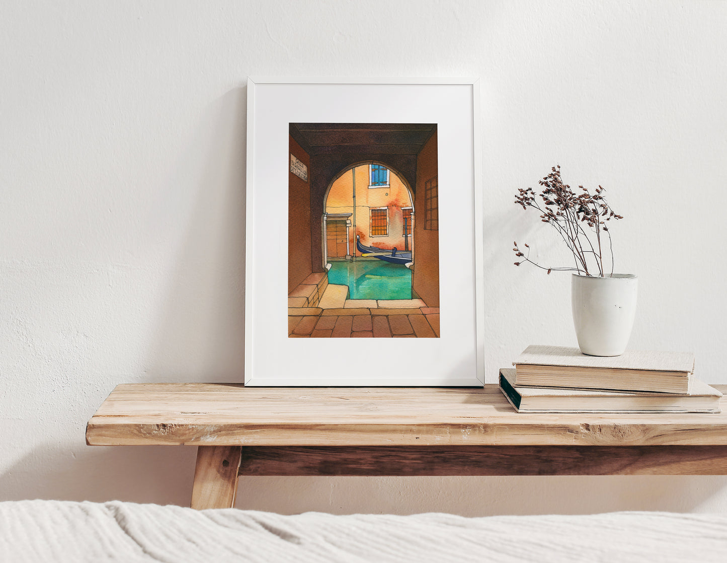 Framed watercolor and ink artwork of a venetian canal scene with a gondola on a wooden shelf with books and a plant.