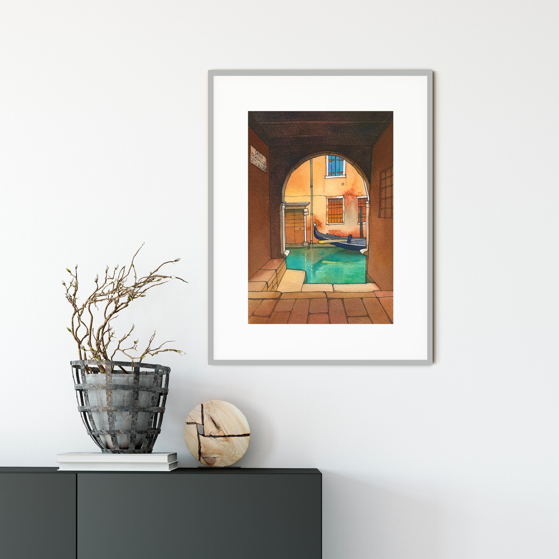 Framed watercolor and ink artwork of a venetian canal scene on a white wall above a black cabinet with decorative items.