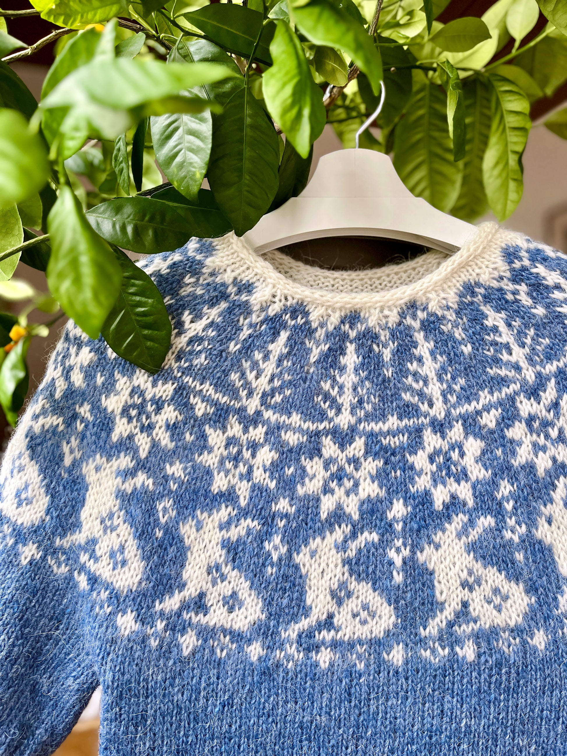 A hand-knitted Icelandic lopapeysa sweater featuring a bunny yoke pattern in blue and white shades, displayed on a hanger against a backdrop of green leaves.