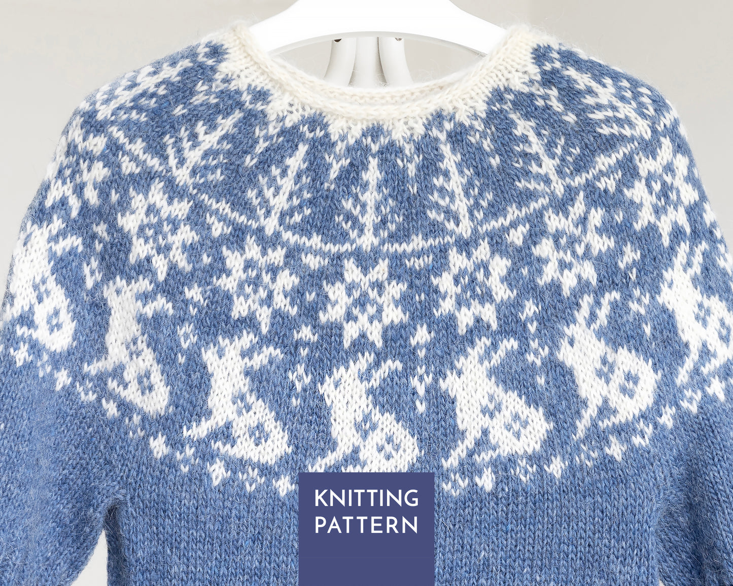 A close-up of a blue and white knitted sweater with a Fair Isle style bunny pattern, hanging on a white hanger against a light gray background
