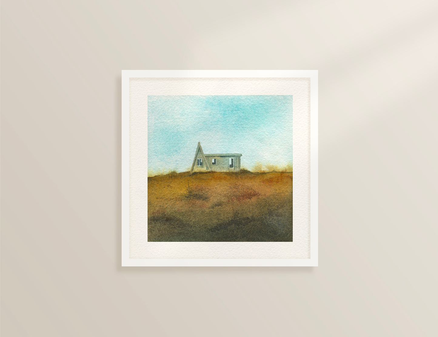 Framed watercolor painting featuring a small A-frame wooden cabin on a grassy hill under a light blue sky in the Westfjords, Iceland