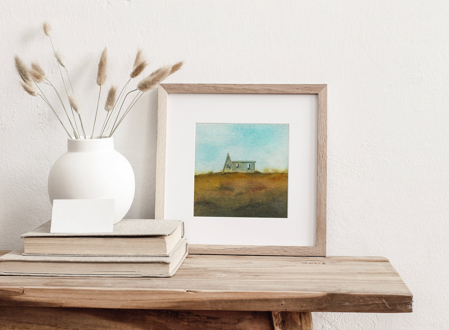 Framed watercolor painting featuring a small A-frame wooden cabin on a grassy hill under a light blue sky in the Westfjords, Iceland, displayed on a wooden table against a white wall.