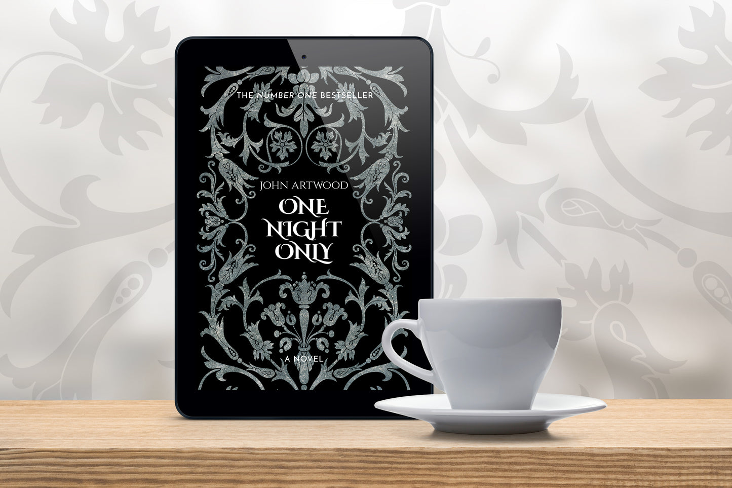 A tablet with a book cover featuring silver baroque ornament on a black background, accompanied with a white cup on a wooden table.