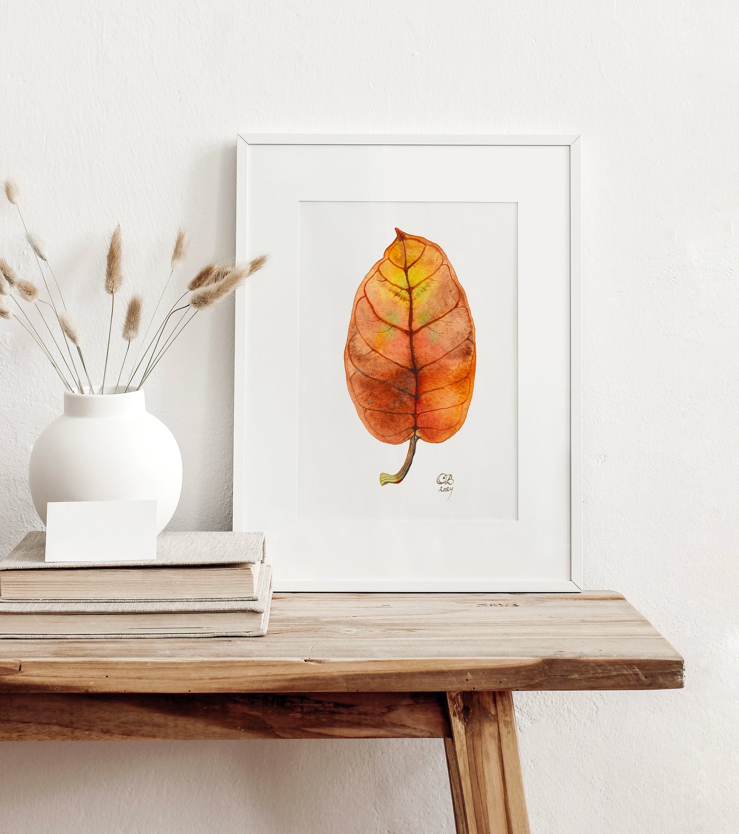 A framed art print featuring a botanical watercolor illustration of a dried autumn leaf, styled on a rustic wooden table next to a white vase.