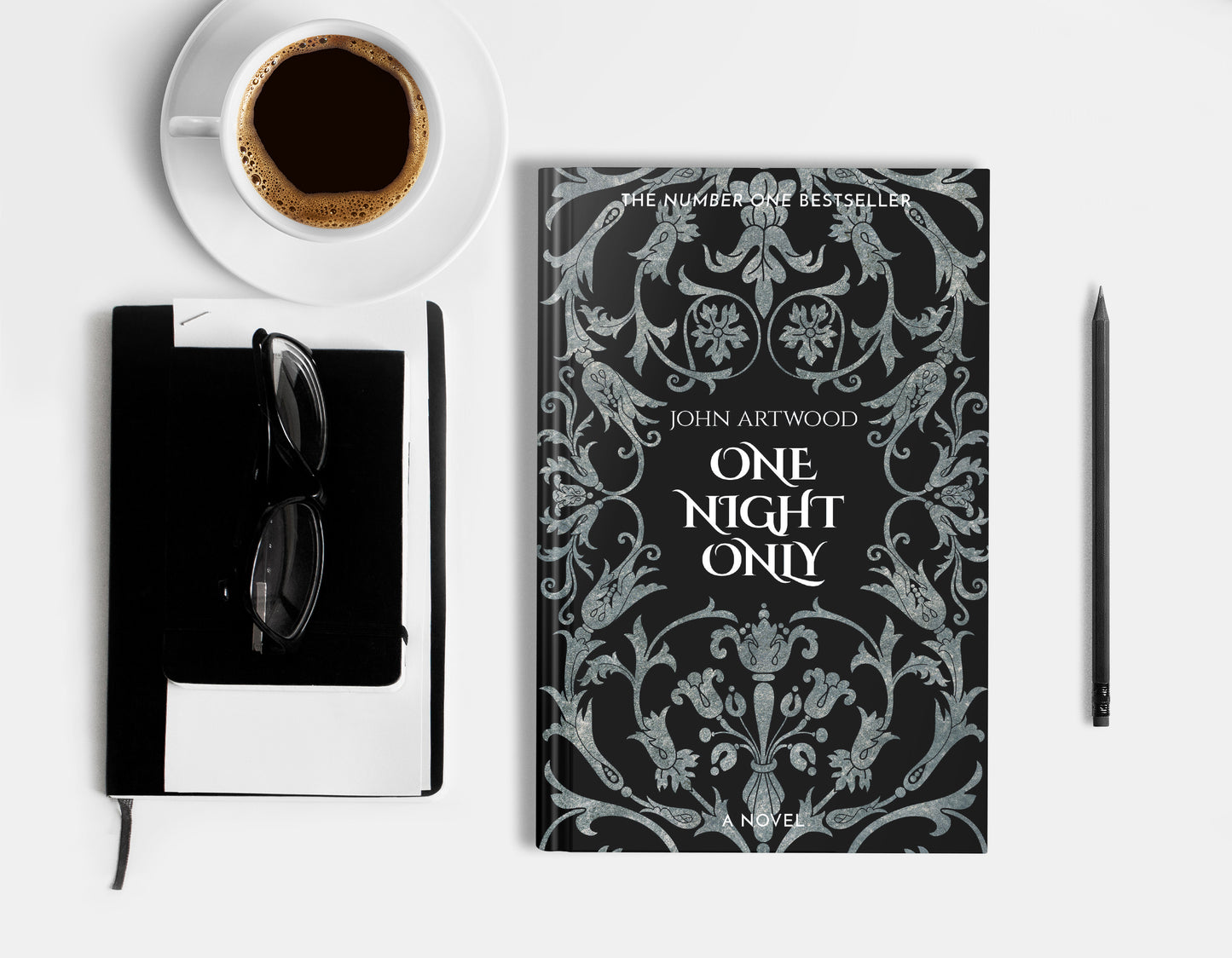 A hardcover book featuring silver baroque ornament on a black background, displaying on a white surface with cup of coffee, a pencil, a notebook and glasses