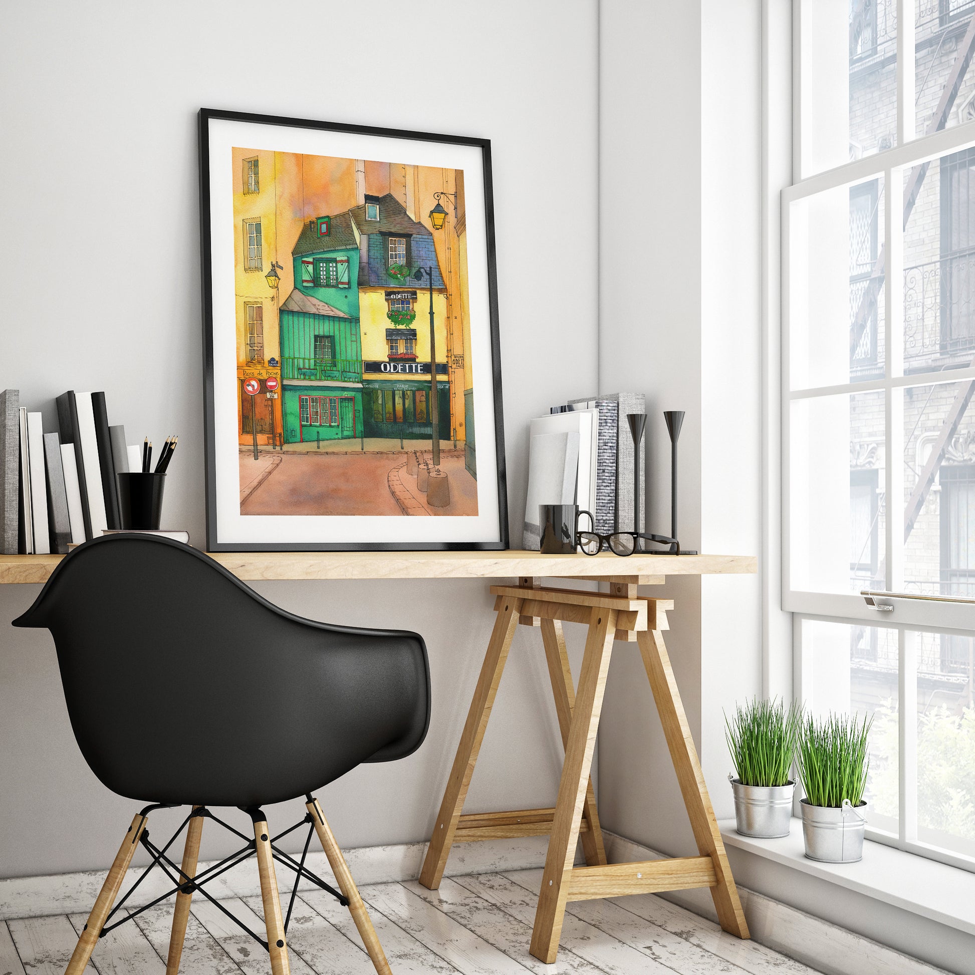 A framed Café Odette in Paris watercolor and ink art print, displayed on a wooden desk in a modern home office setting next to a window.