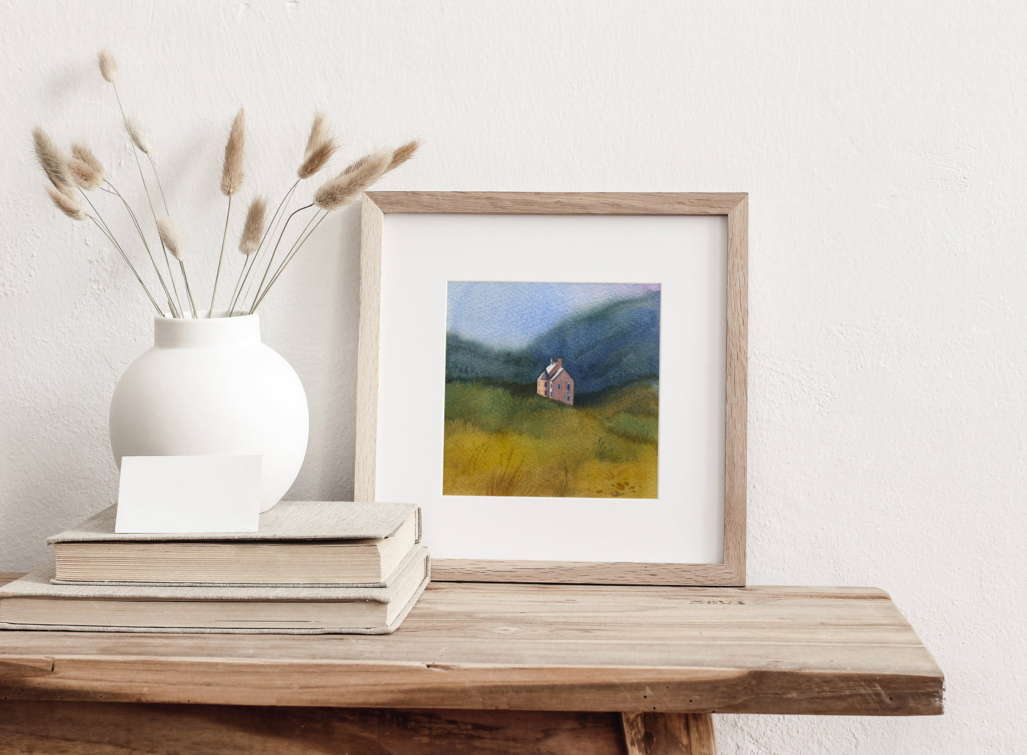 Framed watercolor landscape featuring Culzie Lodge Pink House on Loch Glass in  Scottish Highlands nestled in a grassy field with blue mountains in the background, displayed on a wooden table against a white wall.