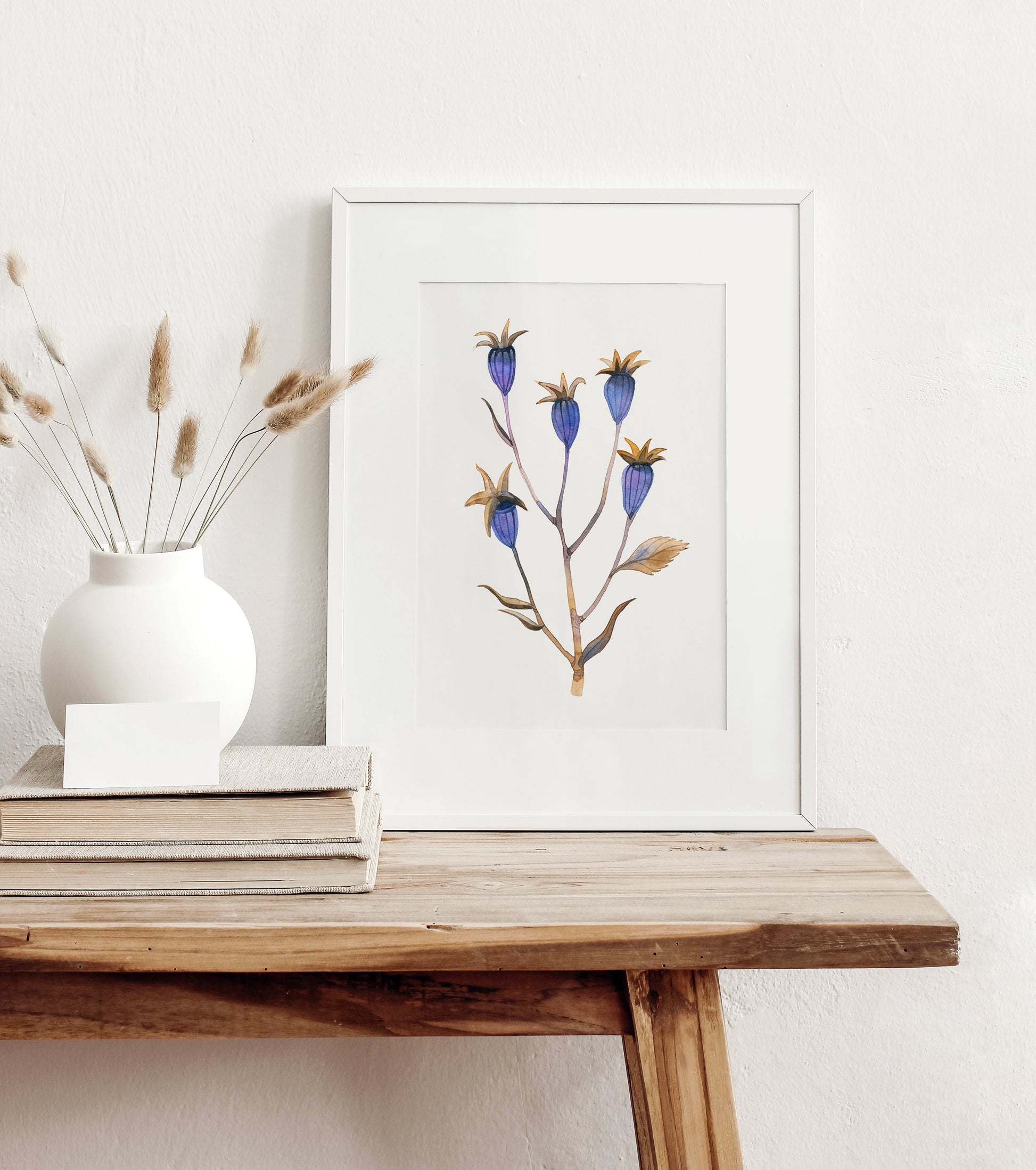 A framed botanical art print featuring dried balloon flower plant, styled on a rustic wooden table next to a white vase with dried grasses and stacked books against a white wall.