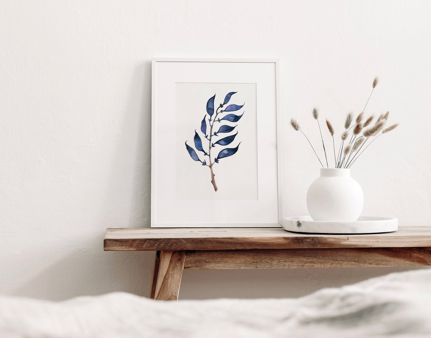 A framed botanical art print featuring a dried longbract wild indigo plant, styled on a wooden table next to a white ceramic vase with dried grasses against a white wall.