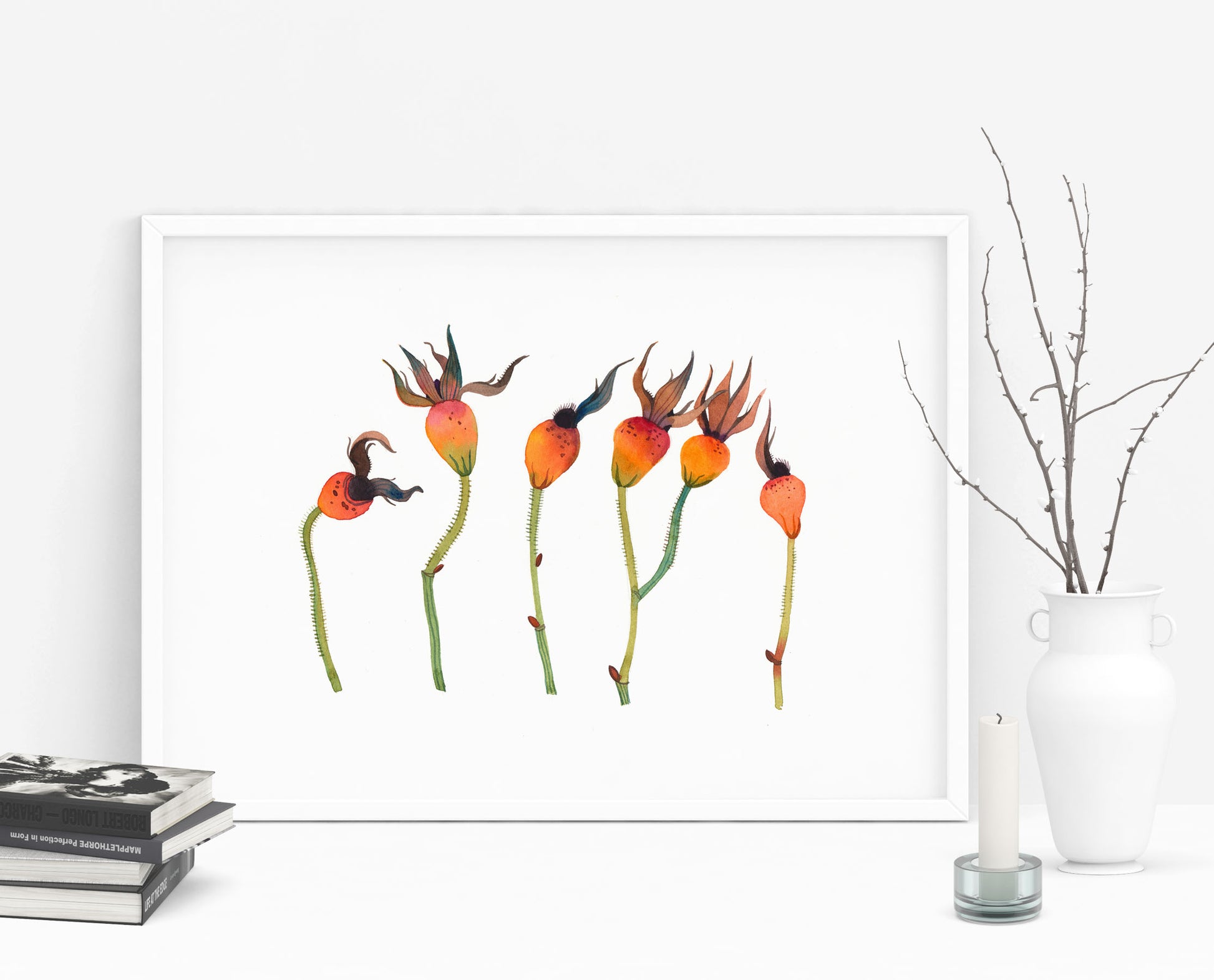 A framed botanical watercolor art print featuring orange dried rose hips with green stems, displayed against a white wall with decorative books, a candle, and a white vase with bare branches.