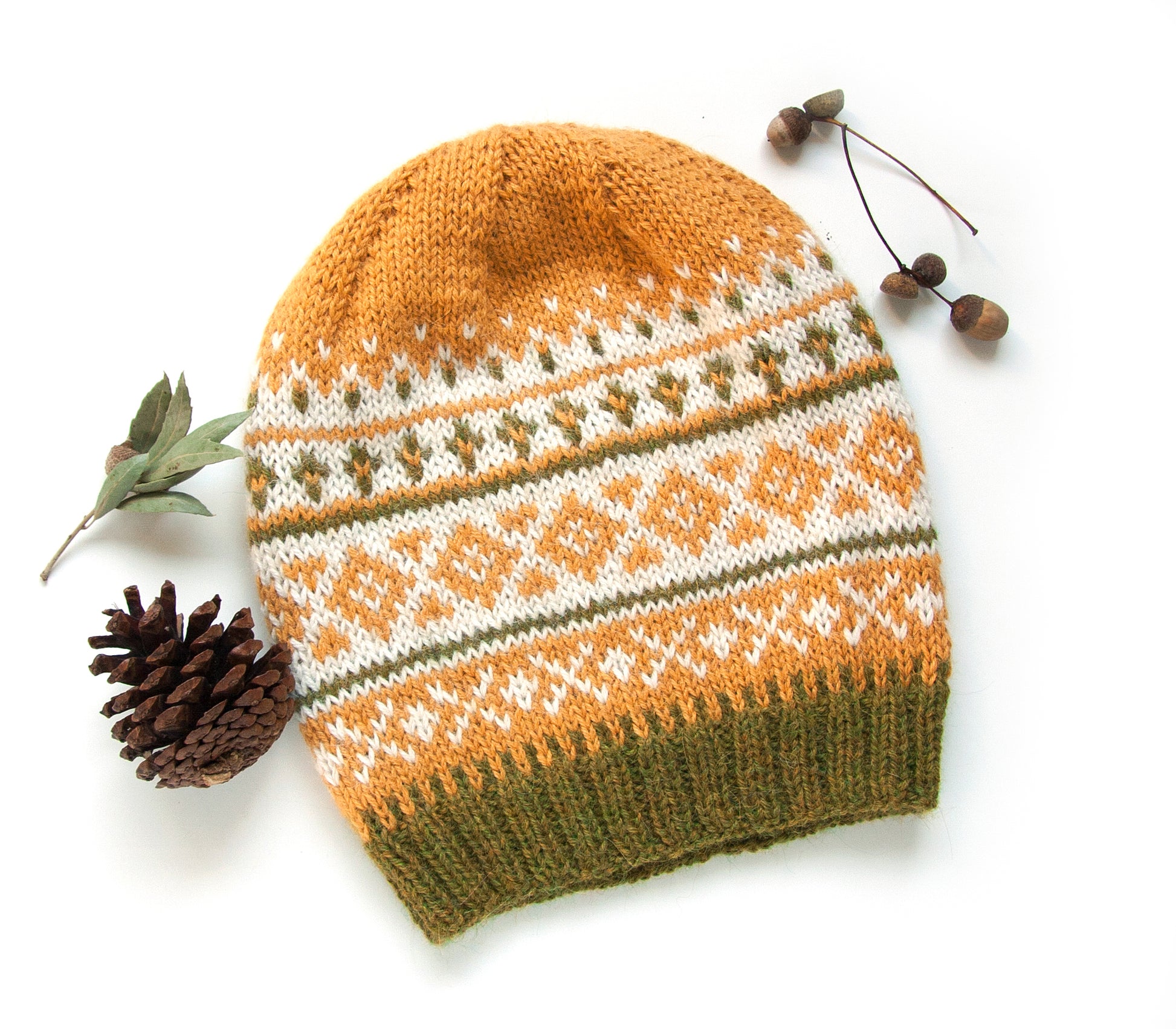Fair Isle hand-knitted alpaca hat in yellow, white, and green, displayed with pine cones on a white background.