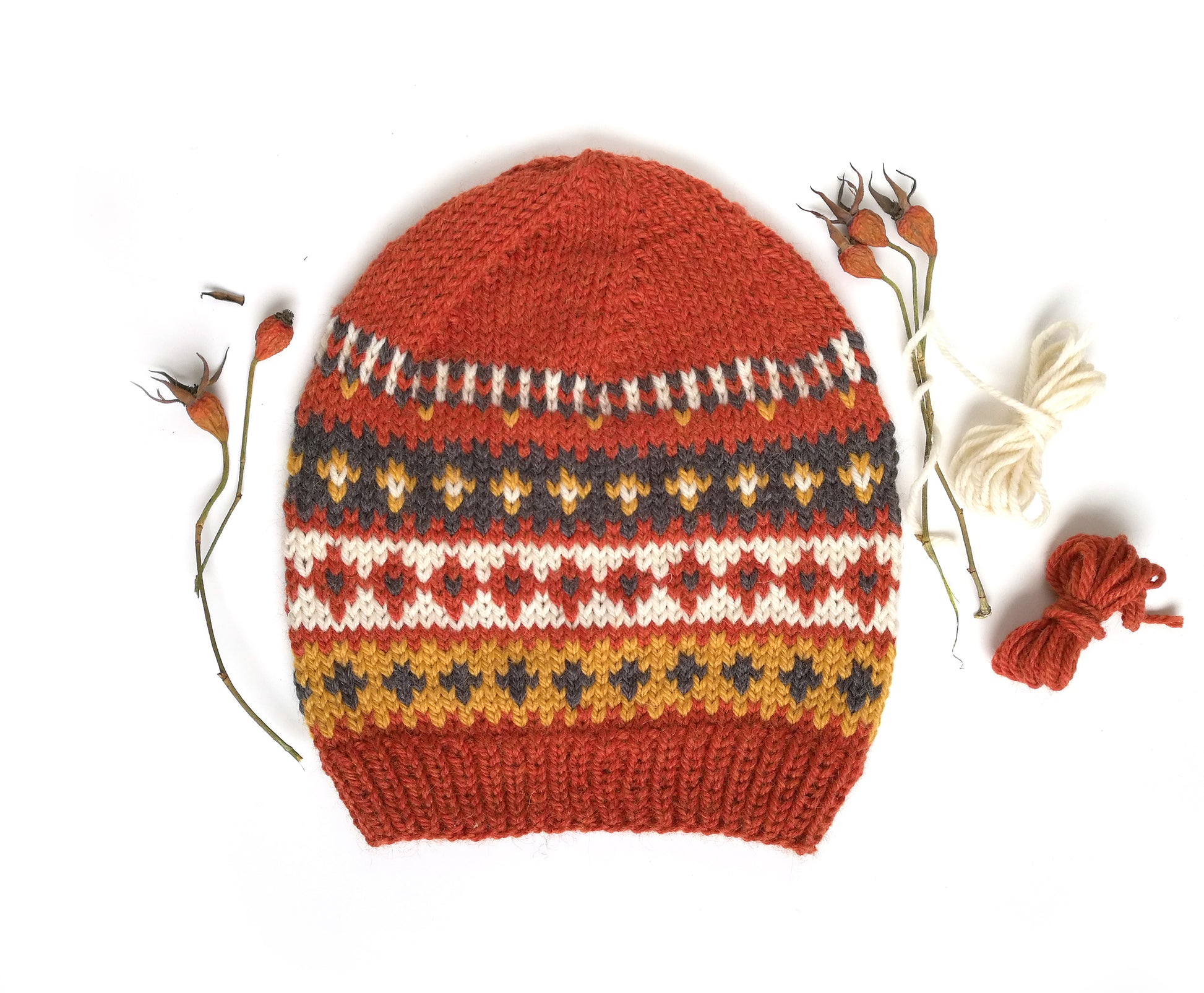 A hand-knitted Fair Isle style hat made of wool and alpaca yarn, with an orange and multicolored geometric pattern, displayed on a white background with decorative dry plants and yarn strands.