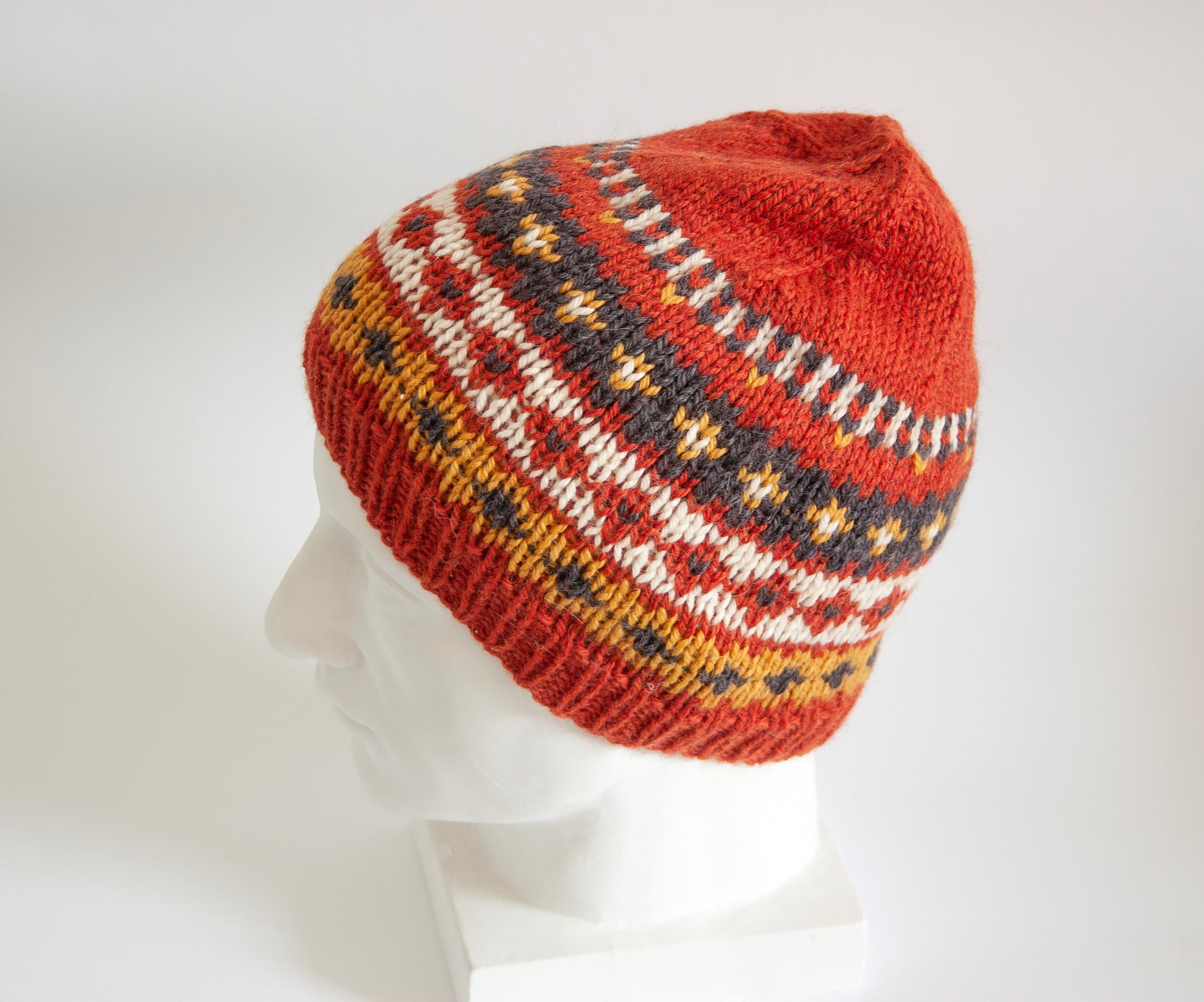A hand-knitted Fair Isle style hat made of wool and alpaca yarn, with an orange and multicolored geometric pattern, displayed on a plaster head mannequin