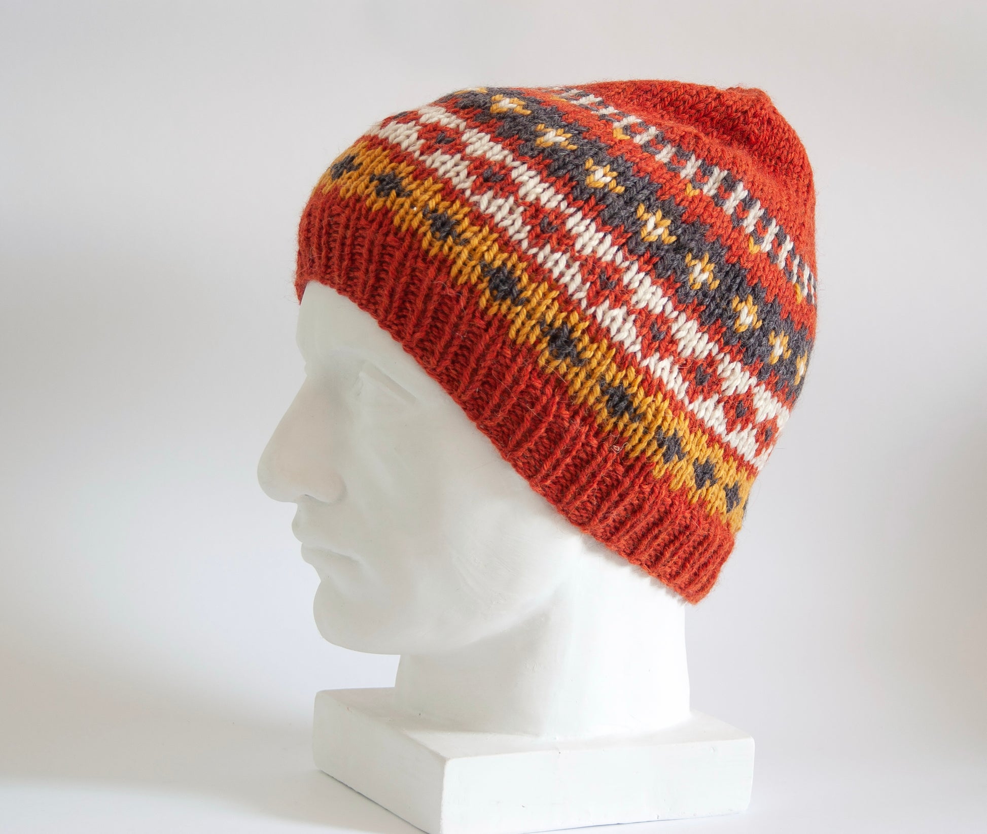 Orange Fair Isle style hat made of wool and alpaca yarn, with multicolored geometric pattern, displayed on a plaster head mannequin