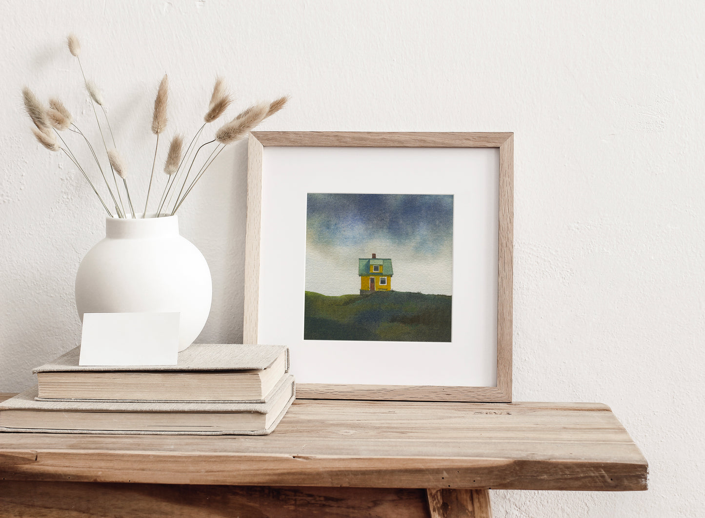 Framed watercolor painting of a small colourfull house on the small car-free Flatey island in the Breiðafjörður fjord, beneath a dramatic Icelandic sky, displayed on a wooden table.