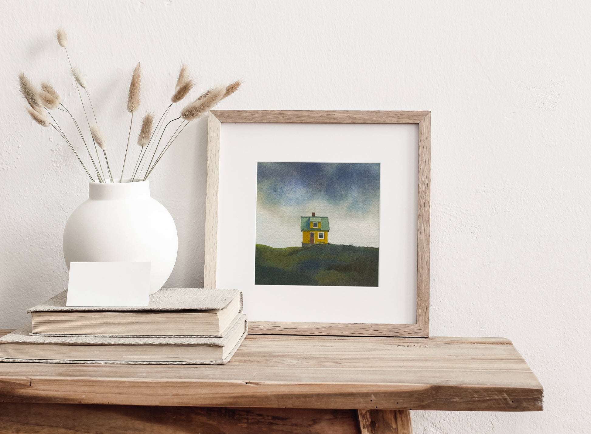 Framed watercolor painting of a small colourfull house on the small car-free Flatey island in the Breiðafjörður fjord, beneath a dramatic Icelandic sky, displayed on a wooden table.