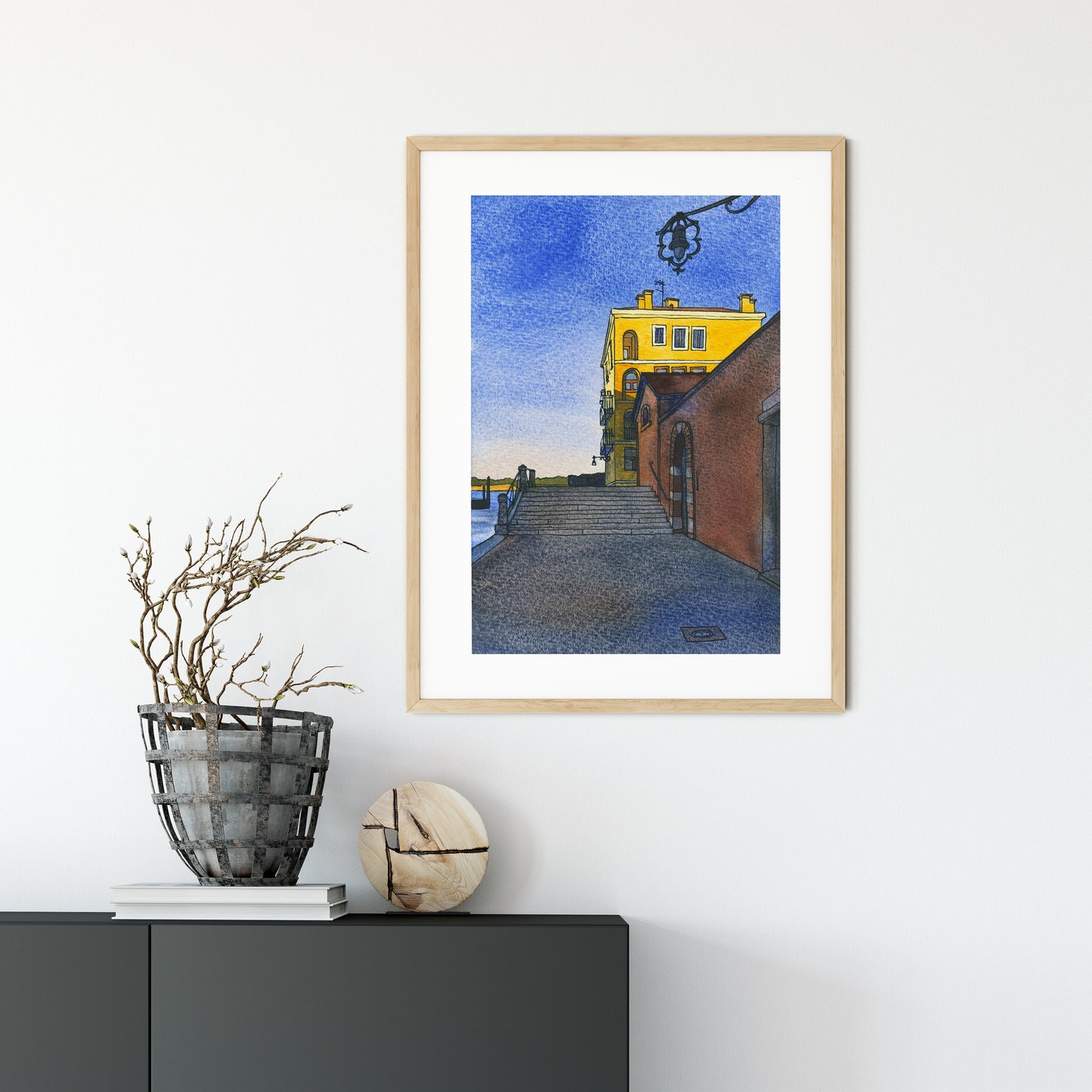 Framed watercolor and ink artwork of a yellow building on Fondamenta Santa Giustina in Venice, on a white wall with decorative items below.