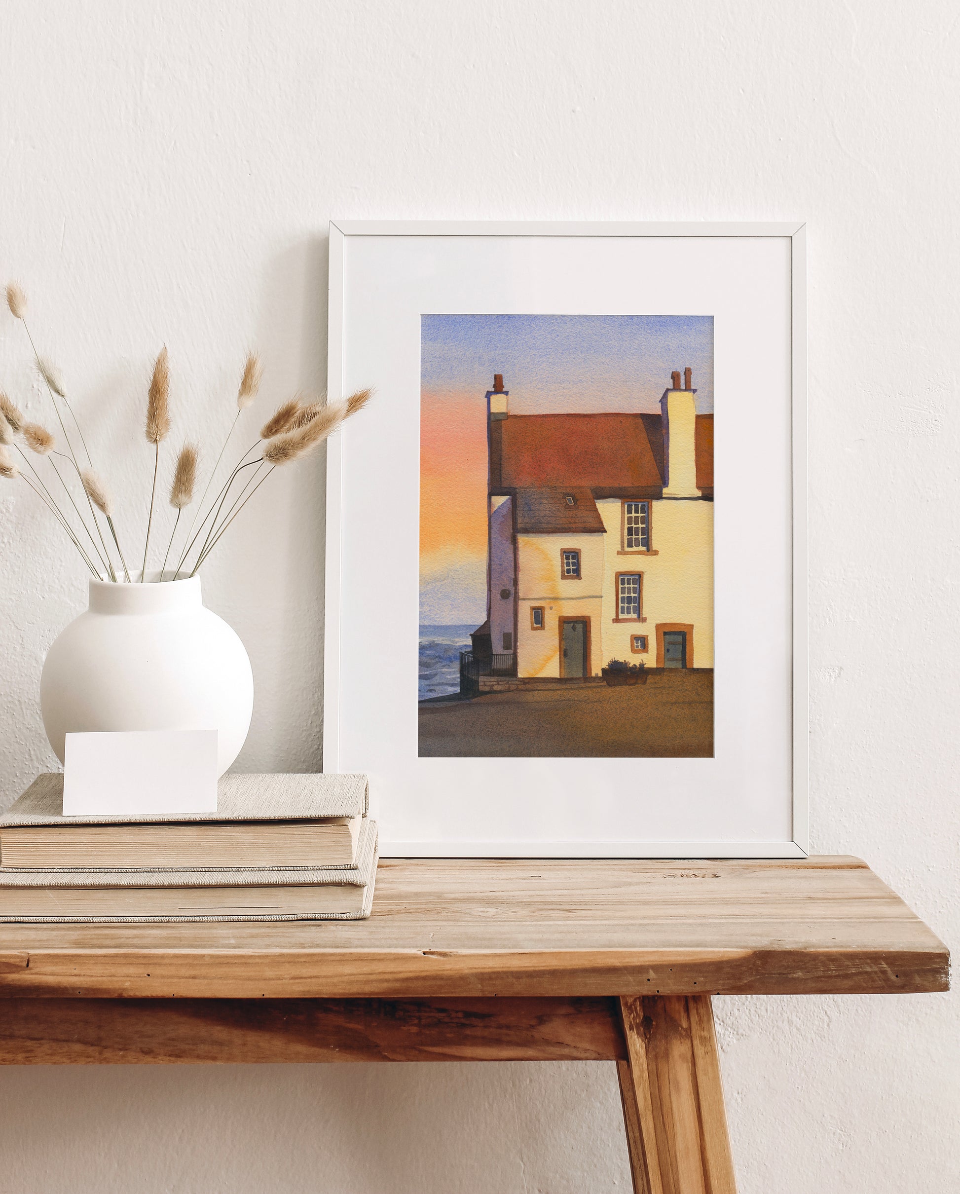 Framed watercolor art print features Glynes House, the coastal cottage in Pittenweem, a picturesque fishing village on Scotland's East Neuk of Fife coast, with its reddish-brown roof, cream walls, chimneys, and a pale sky with a soft sunset glow, displayed on a wooden table with decorative items.