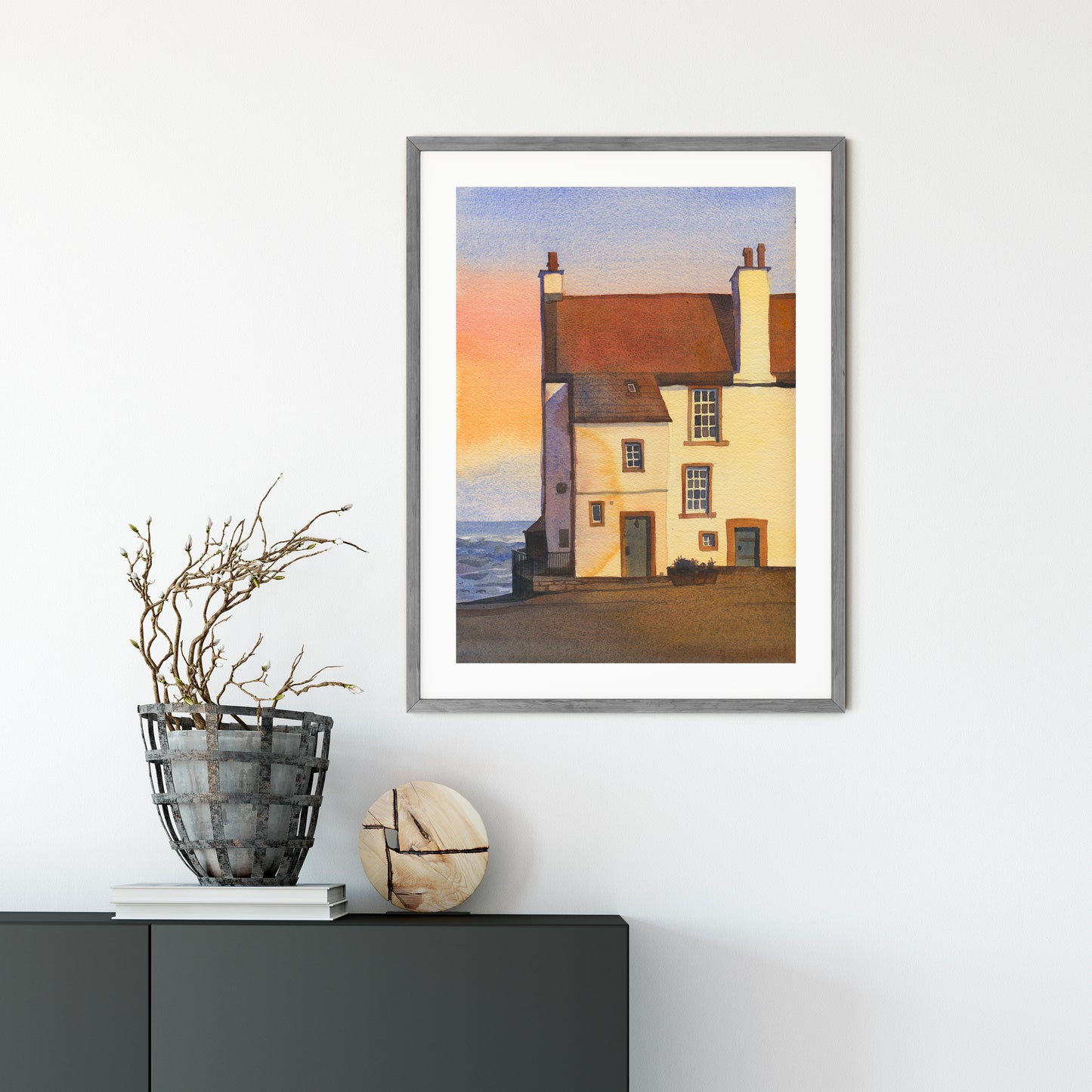 Framed watercolor artwork features Glynes House, the coastal cottage in Pittenweem, a picturesque fishing village on Scotland's East Neuk of Fife coast, with its reddish-brown roof, cream walls, chimneys, and a pale sky with a soft sunset glow.