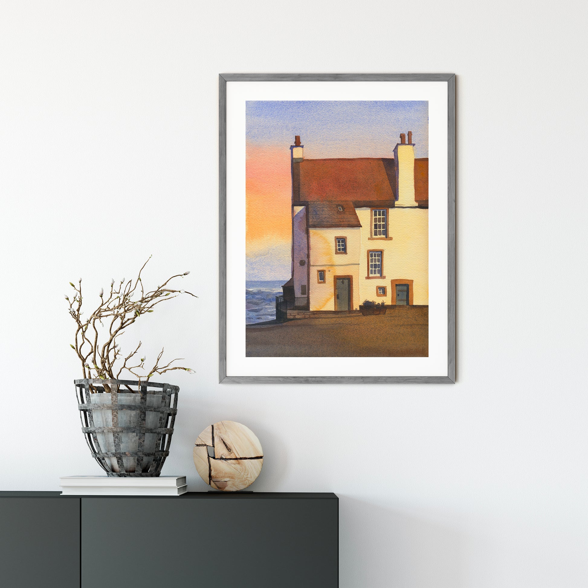 Framed watercolor artwork features Glynes House, the coastal cottage in Pittenweem, a picturesque fishing village on Scotland's East Neuk of Fife coast, with its reddish-brown roof, cream walls, chimneys, and a pale sky with a soft sunset glow.