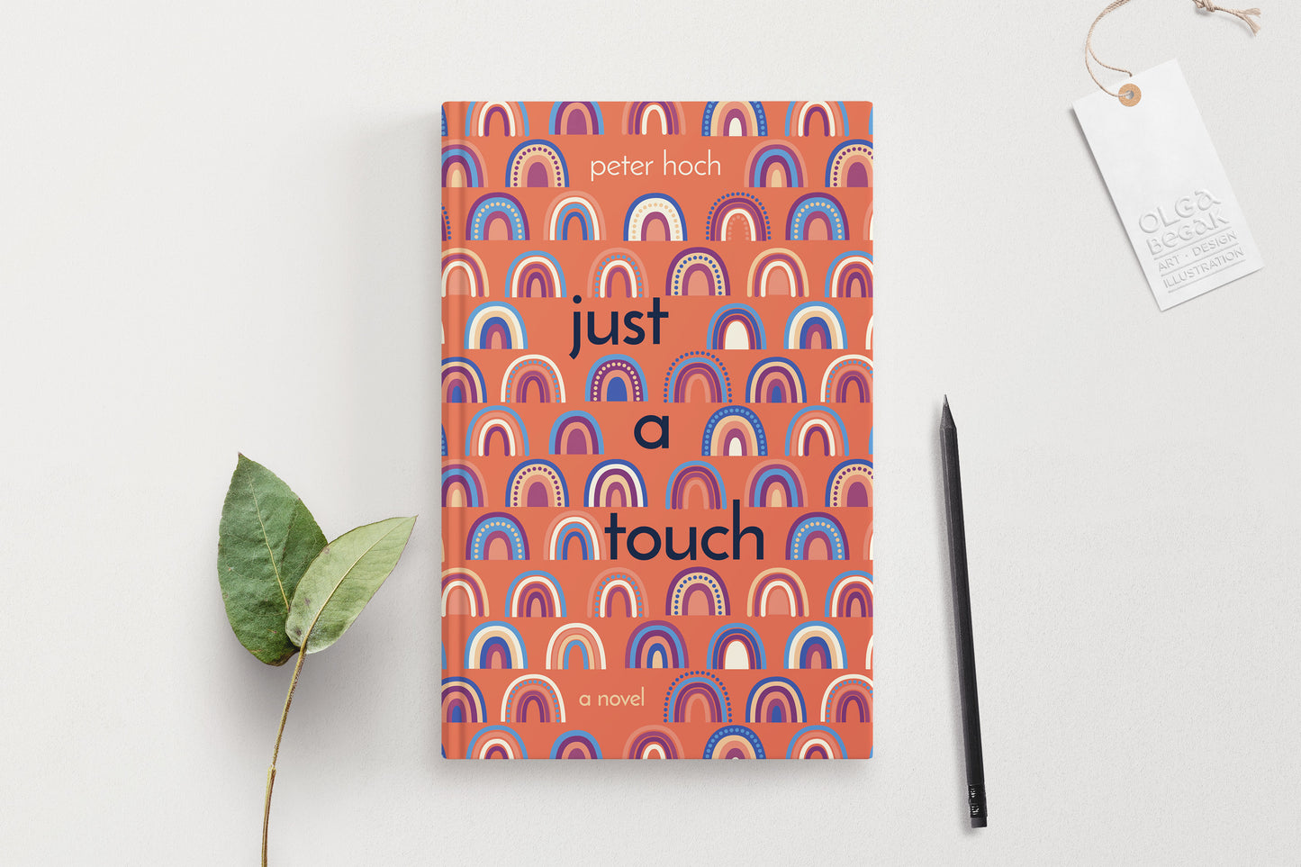 A hardcover book featuring colorful rainbow ornaments on a coral background, displaying on a white surface with pencil and eucalyptus branch.