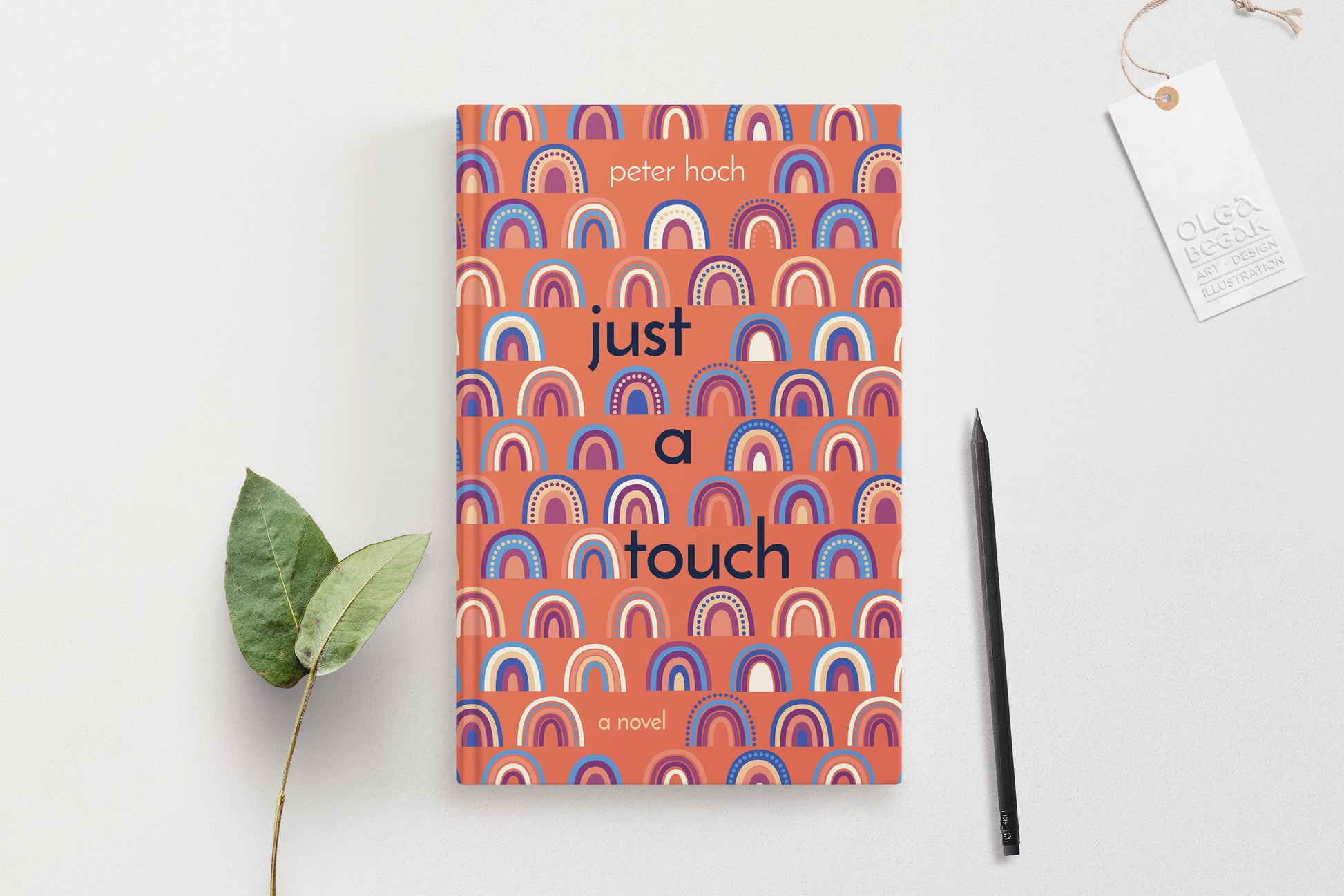 A hardcover book featuring colorful rainbow ornaments on a coral background, displaying on a white surface with pencil and eucalyptus branch.