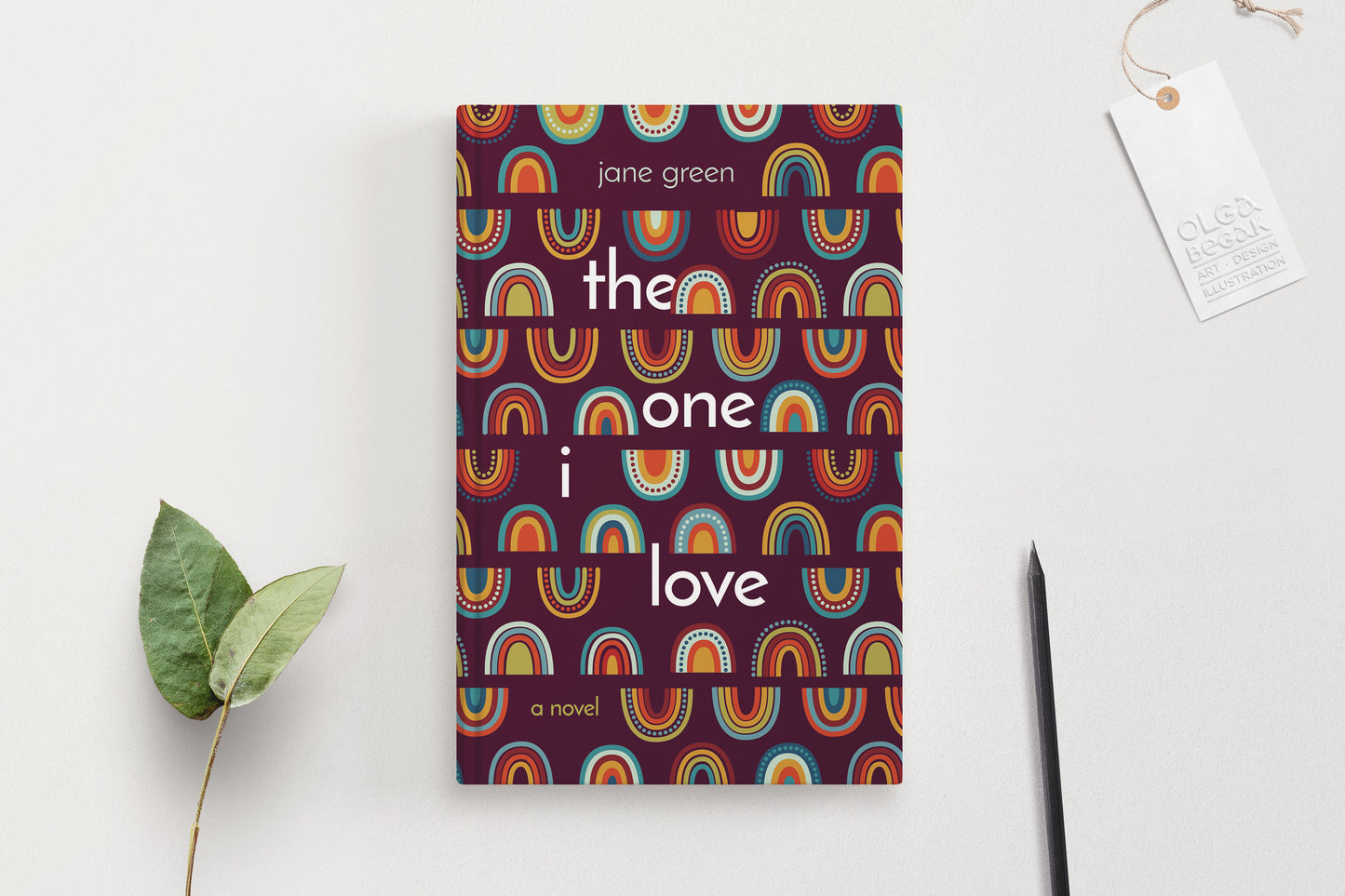 A hardcover book featuring colorful rainbow ornaments on a purple background, displaying on a white surface with pencil and eucalyptus branch.