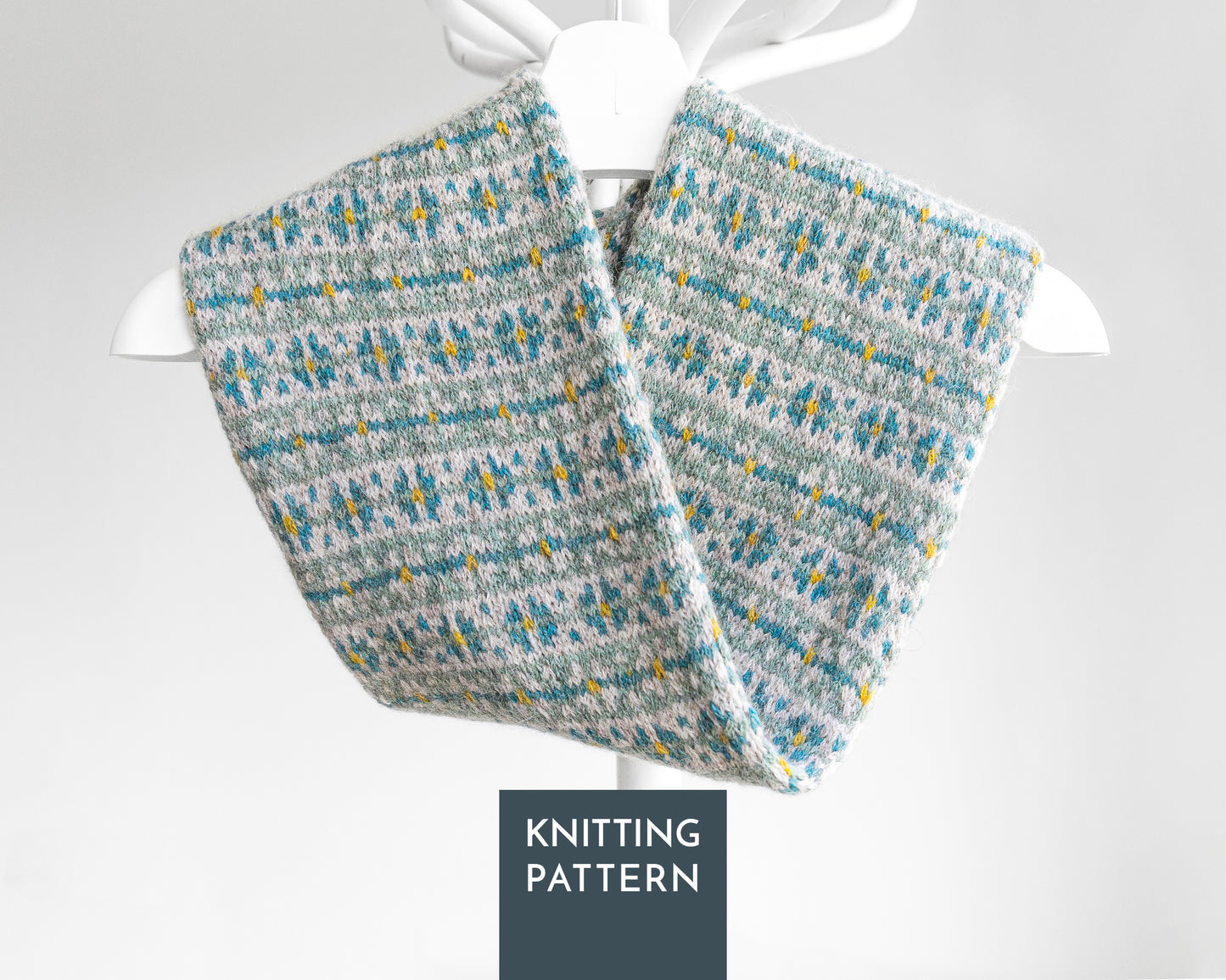 A knitted twisted cowl scarf with a gray, teal blue, and yellow Fair Isle pattern. A dark gray label at the bottom reads 'KNITTING PATTERN'.