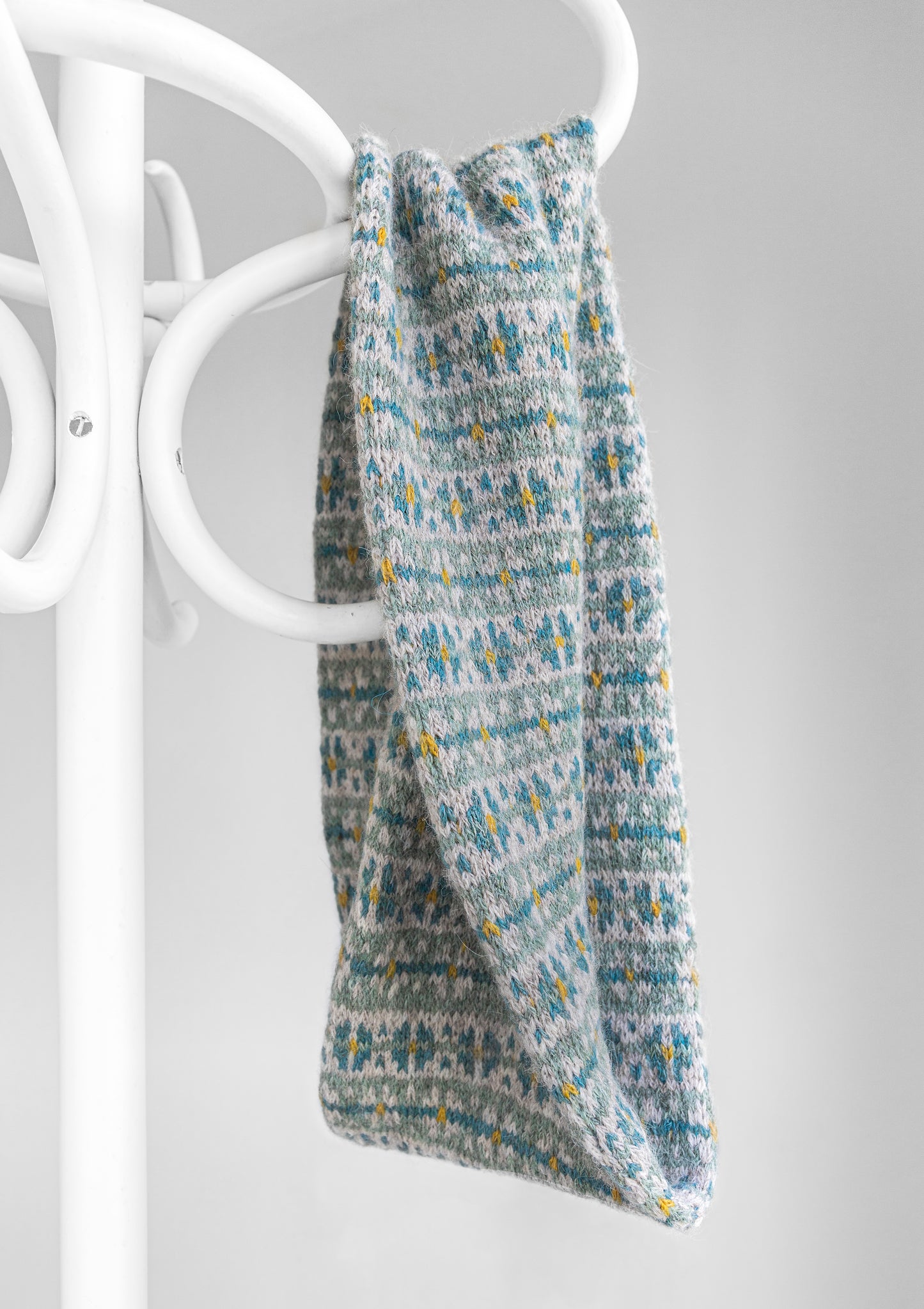 A knitted twisted cowl scarf with a gray, teal blue, and yellow Fair Isle pattern, draped over a white coat rack against a light gray background.