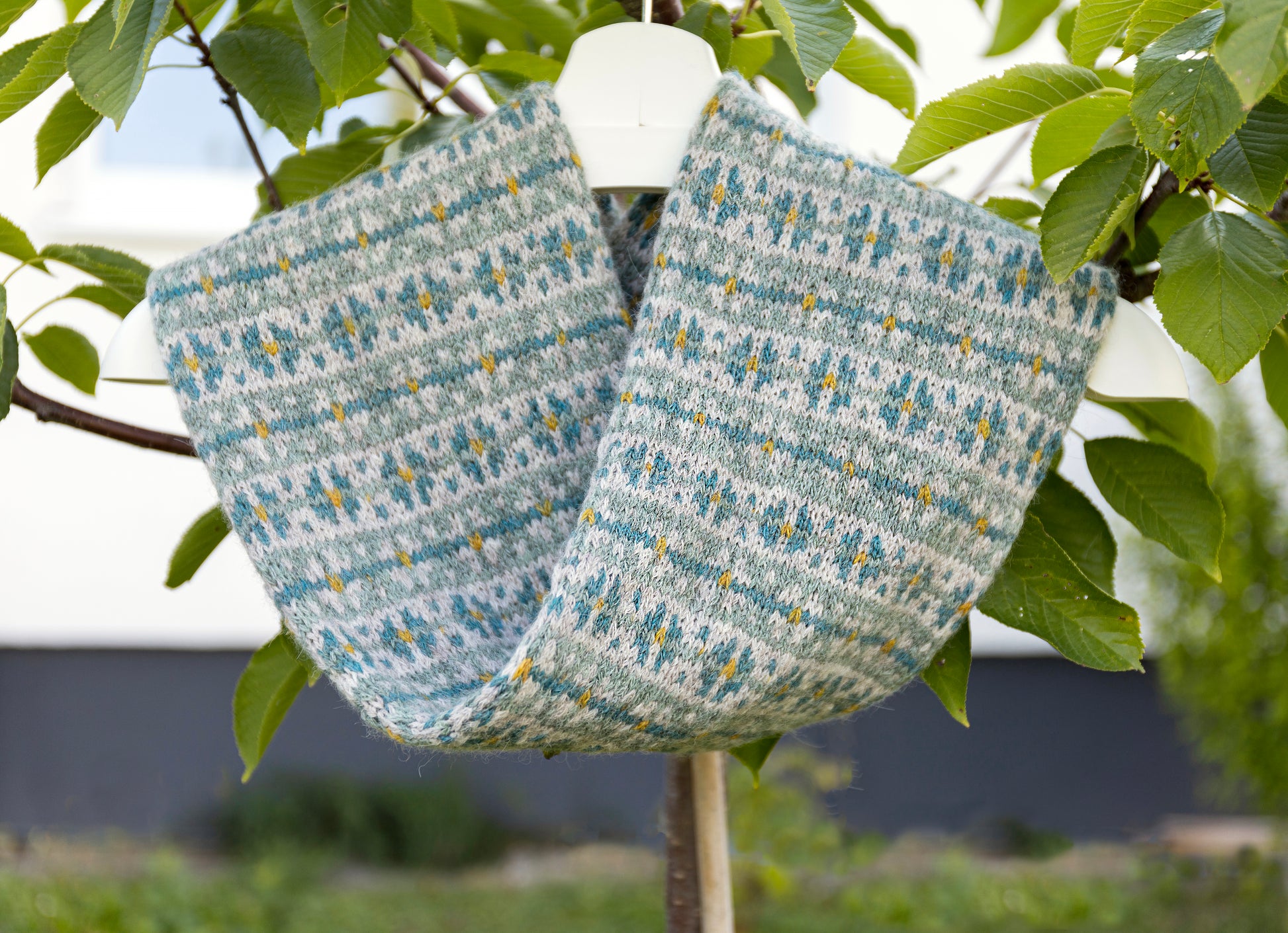 A knitted twisted cowl scarf with a gray, teal blue, and yellow Fair Isle pattern, hanging from a white hanger on a tree branch outdoors.