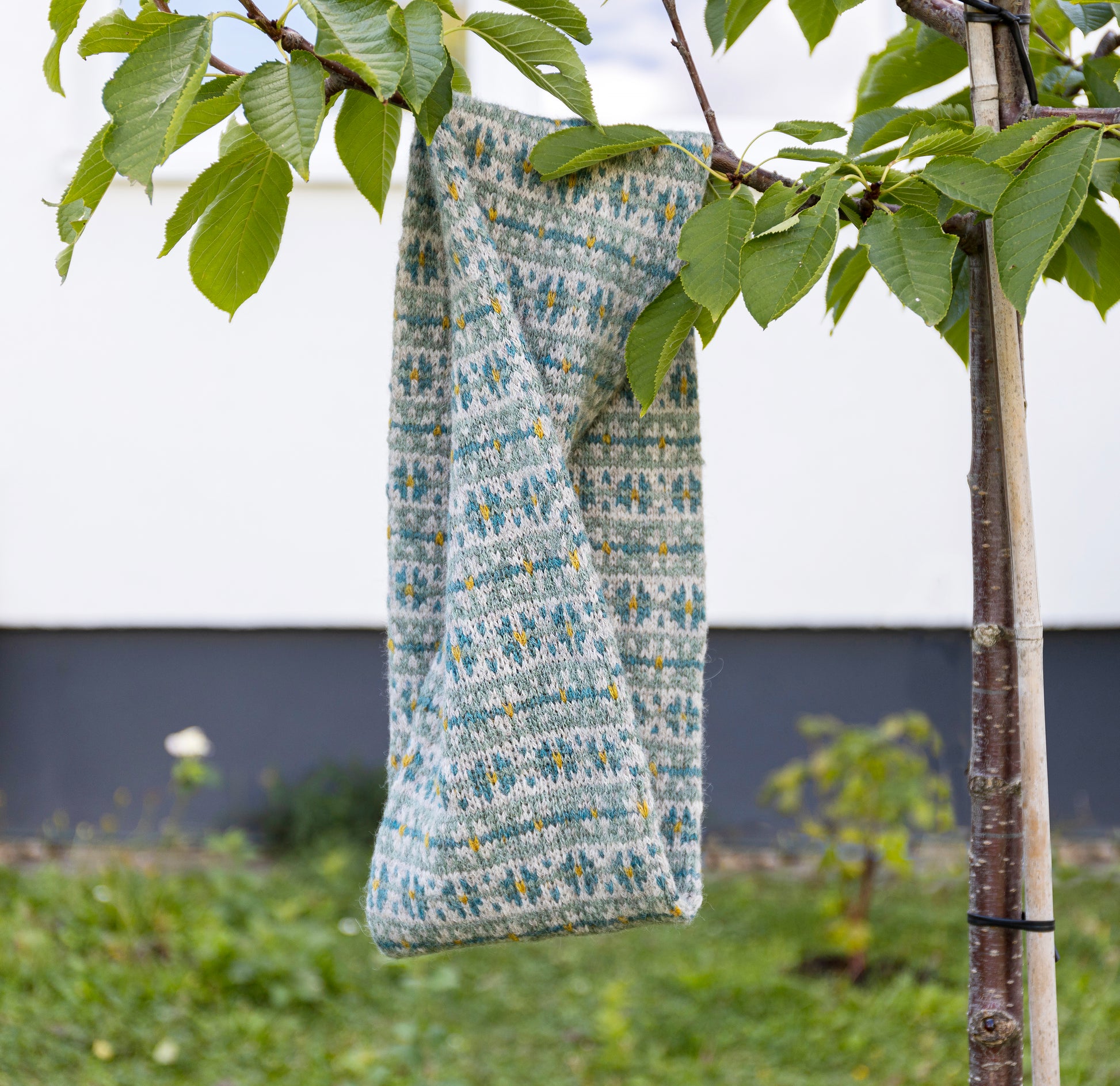 A knitted twisted cowl scarf with a gray, teal blue, and yellow Fair Isle pattern hangs from a tree branch outdoors, with green leaves framing the top of the shot.