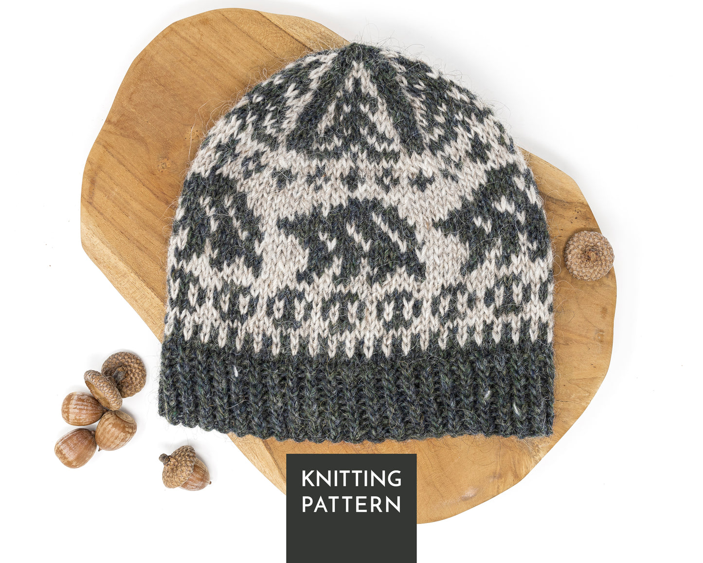 A knitted beanie hat with a dark gray-green and light beige Fair Isle style pattern, featuring bears, displayed on a wooden board with acorns. A black label at the bottom reads 'KNITTING PATTERN'.