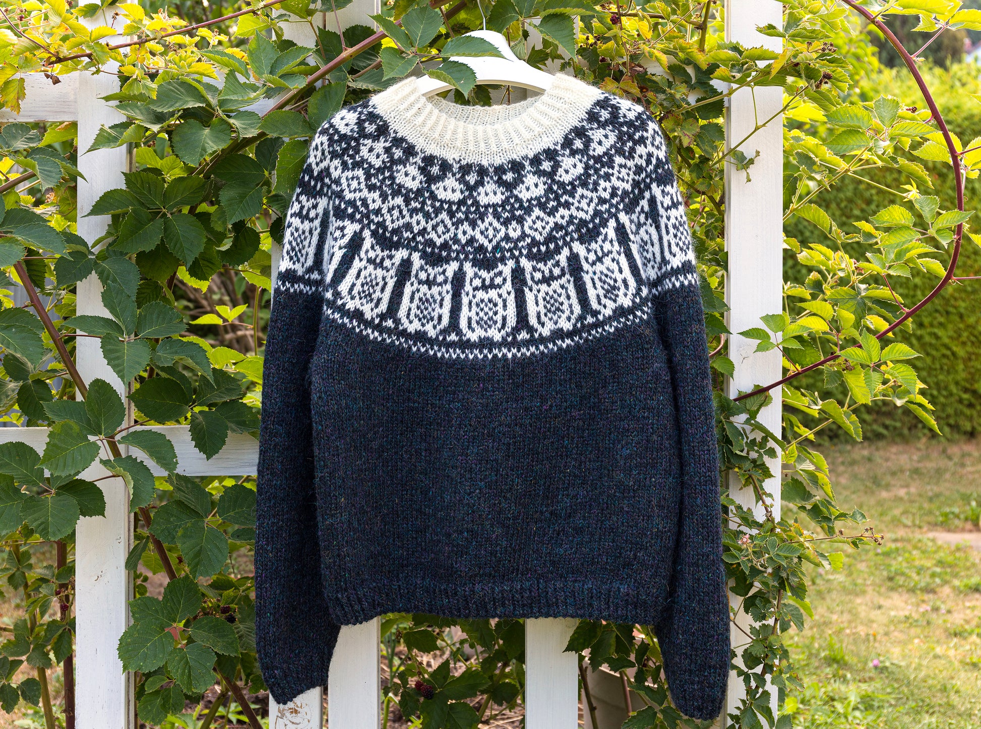 A black and white knit sweater with Nattuggla Nigh Owl pattern, hanging on a white hanger outdoors against a white fence and green foliage.