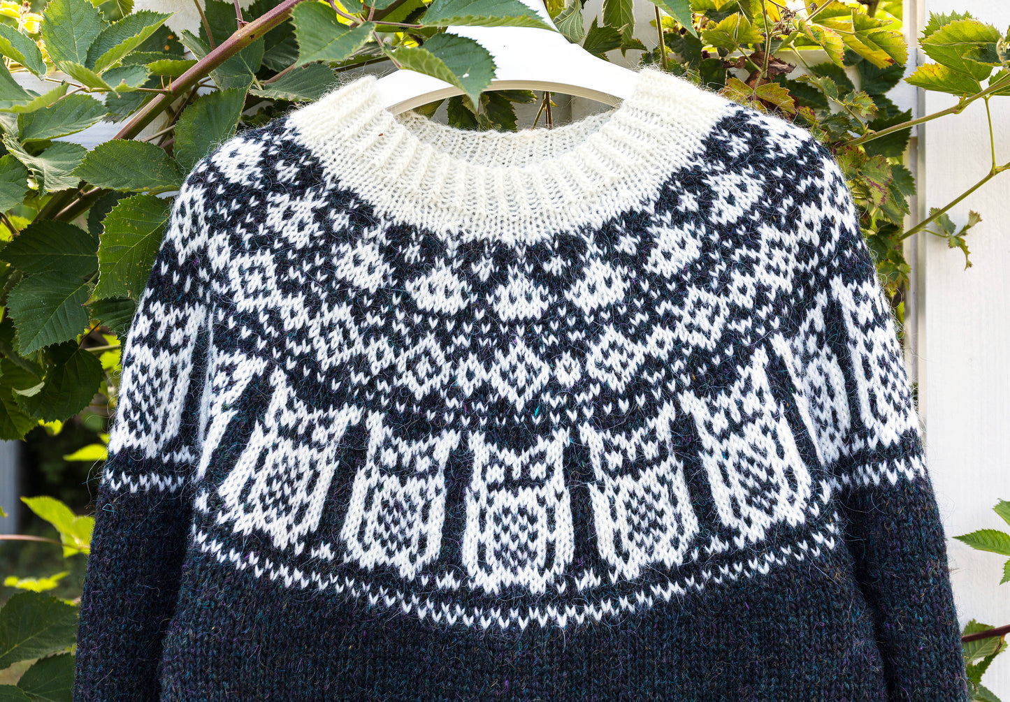 A black and white knitted Icelandic sweater with a traditional yoke in Nattuggla Nigh Owl pattern, hanging on a white hanger against a background of green leaves.