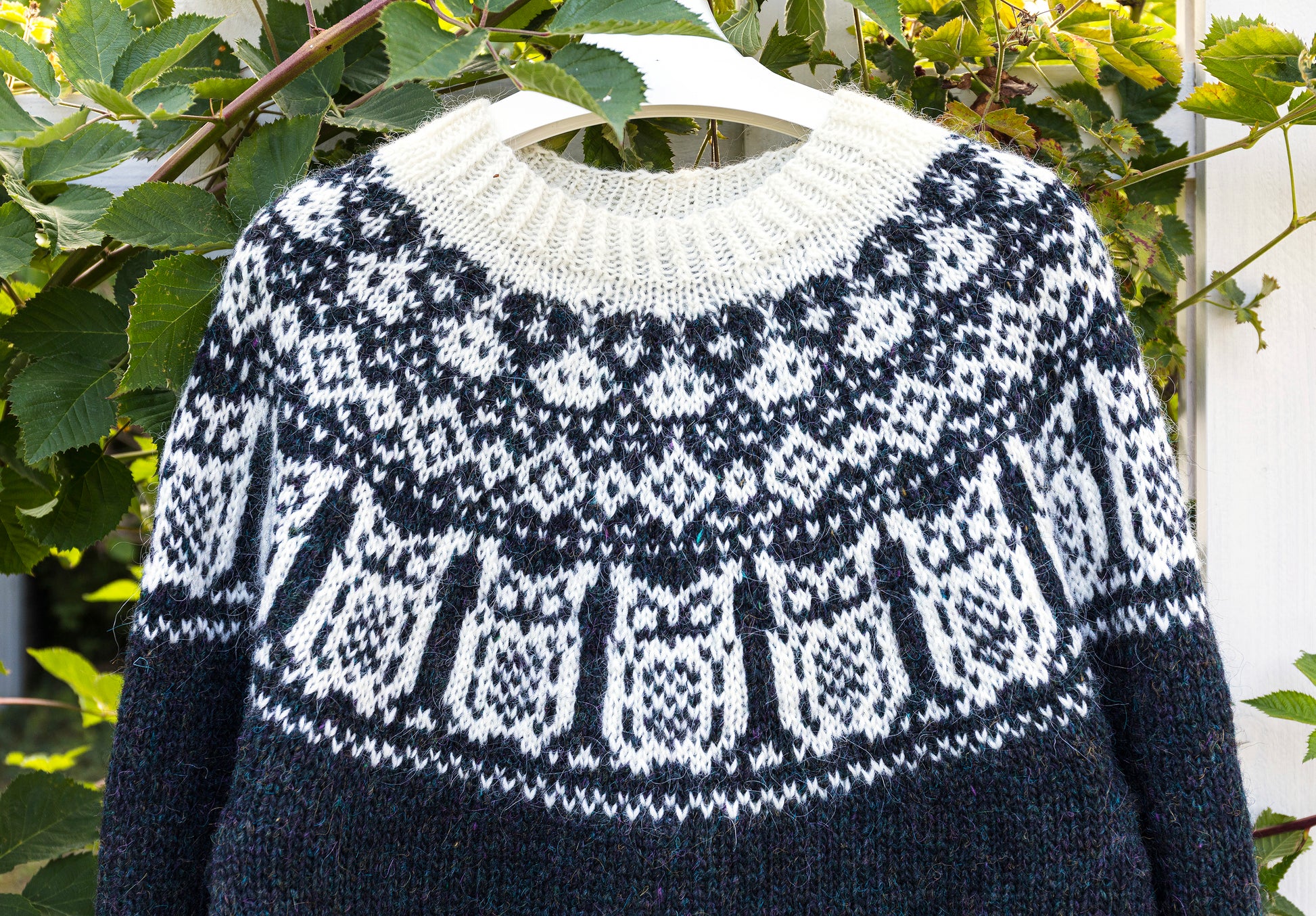 A black and white knitted Icelandic sweater with a traditional yoke in Nattuggla Nigh Owl pattern, hanging on a white hanger against a background of green leaves.