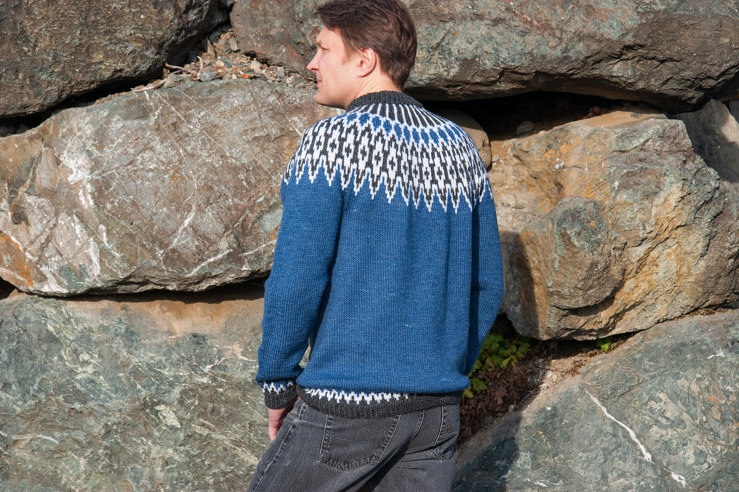 A man wearing a blue knit Icelandic lopapeysa sweater with a white and black geometric pattern across the yoke, standing outdoors in front of large natural rocks.