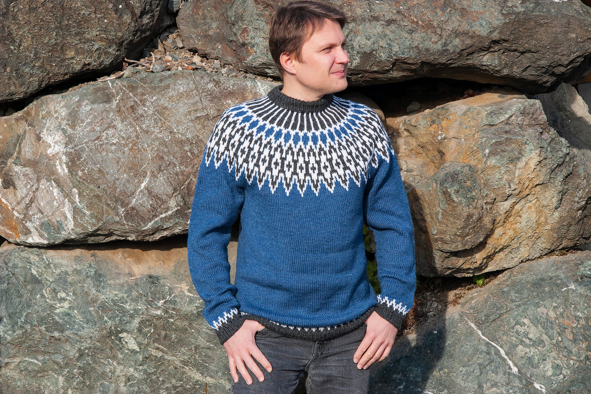 A man wearing a blue knit Icelandic sweater with a white and black geometric Fair Isle yoke pattern, standing outdoors against a background of large natural rocks.