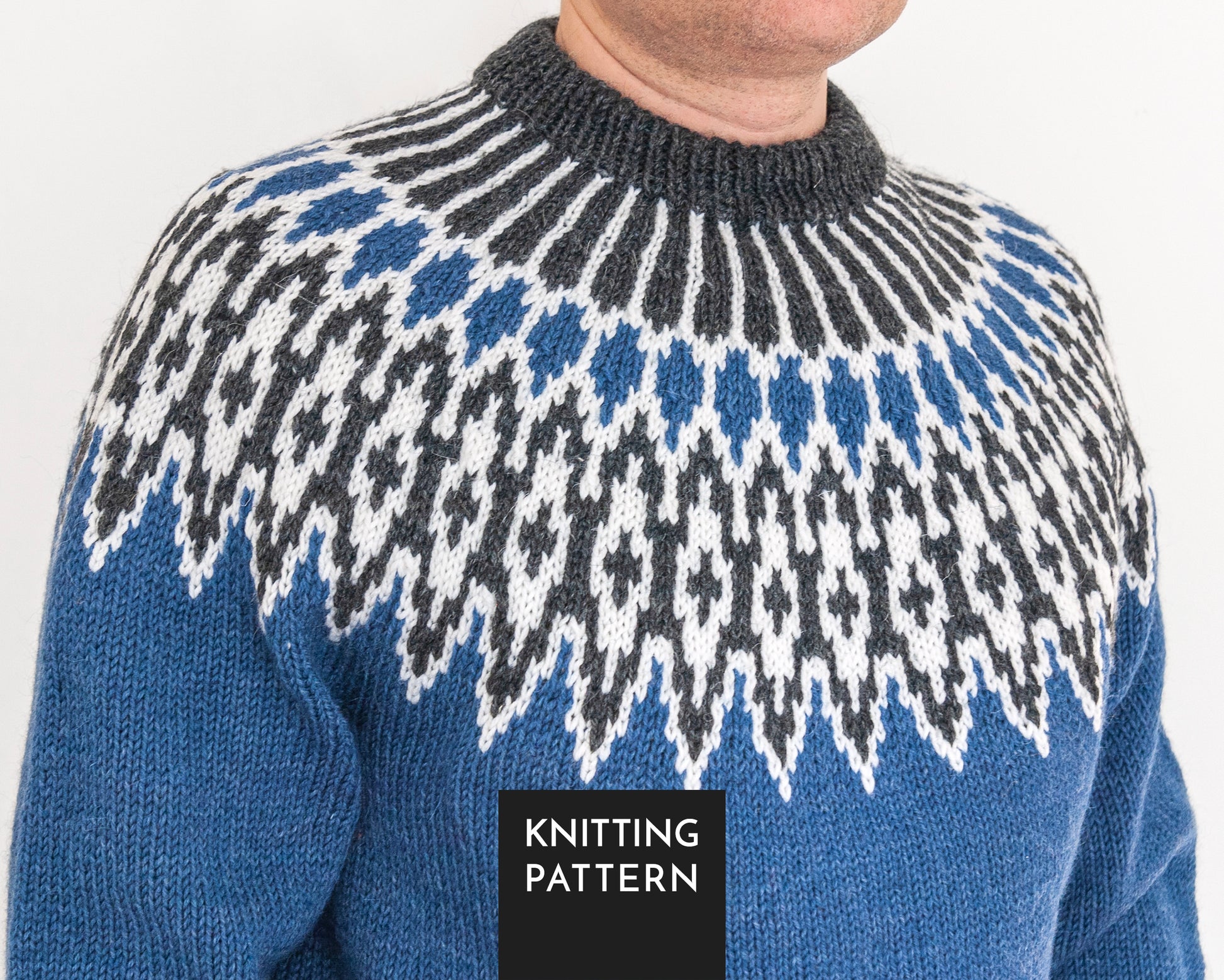 A close-up of a knitted lopapeysa sweater worn by a person. The sweater has a blue base with a large Fair Isle style yoke pattern in white and dark gray, and a dark gray ribbed crew neck.